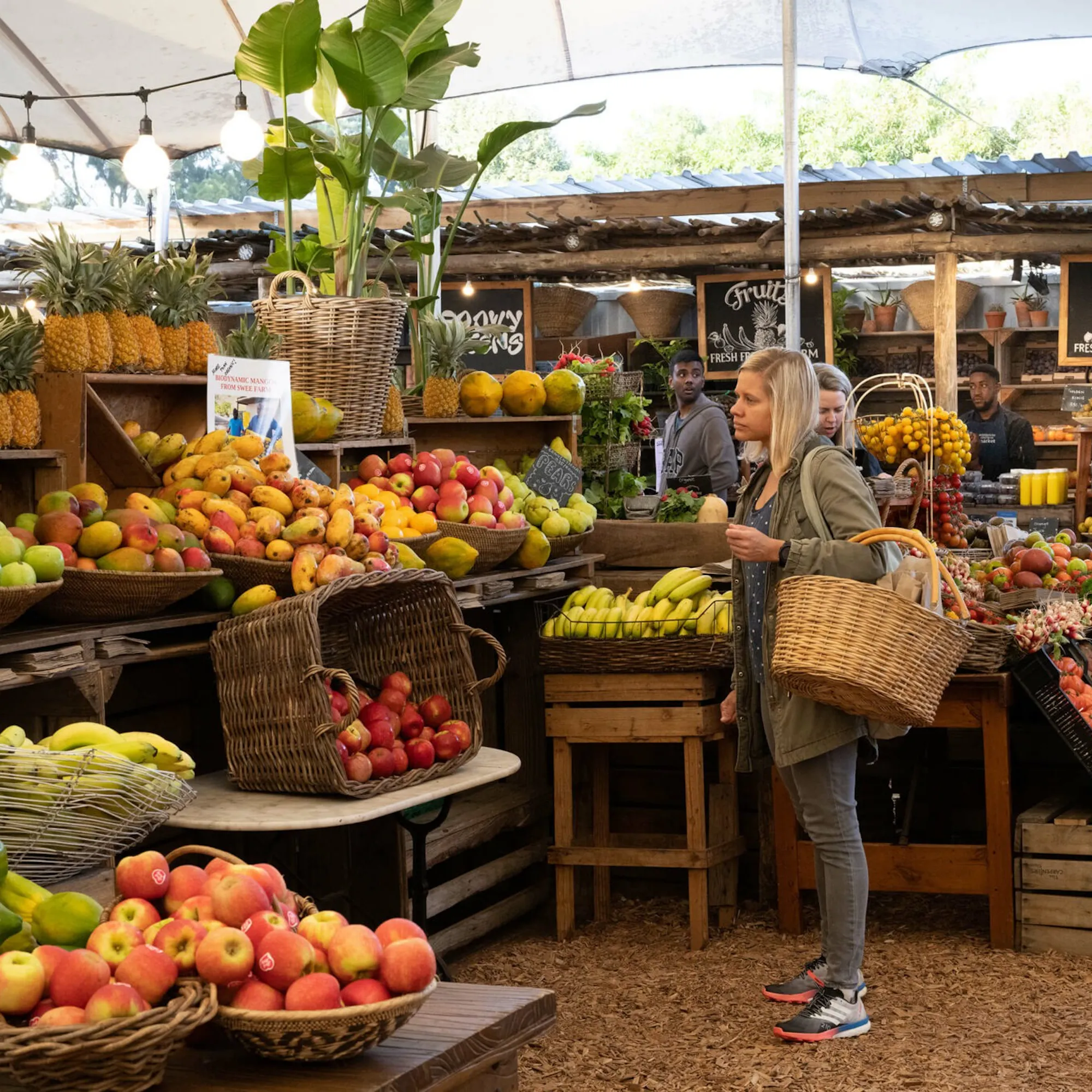 A shopper with a woven basket stands among bright fruit and vegetable displays at Oranjezicht City Farm Market.