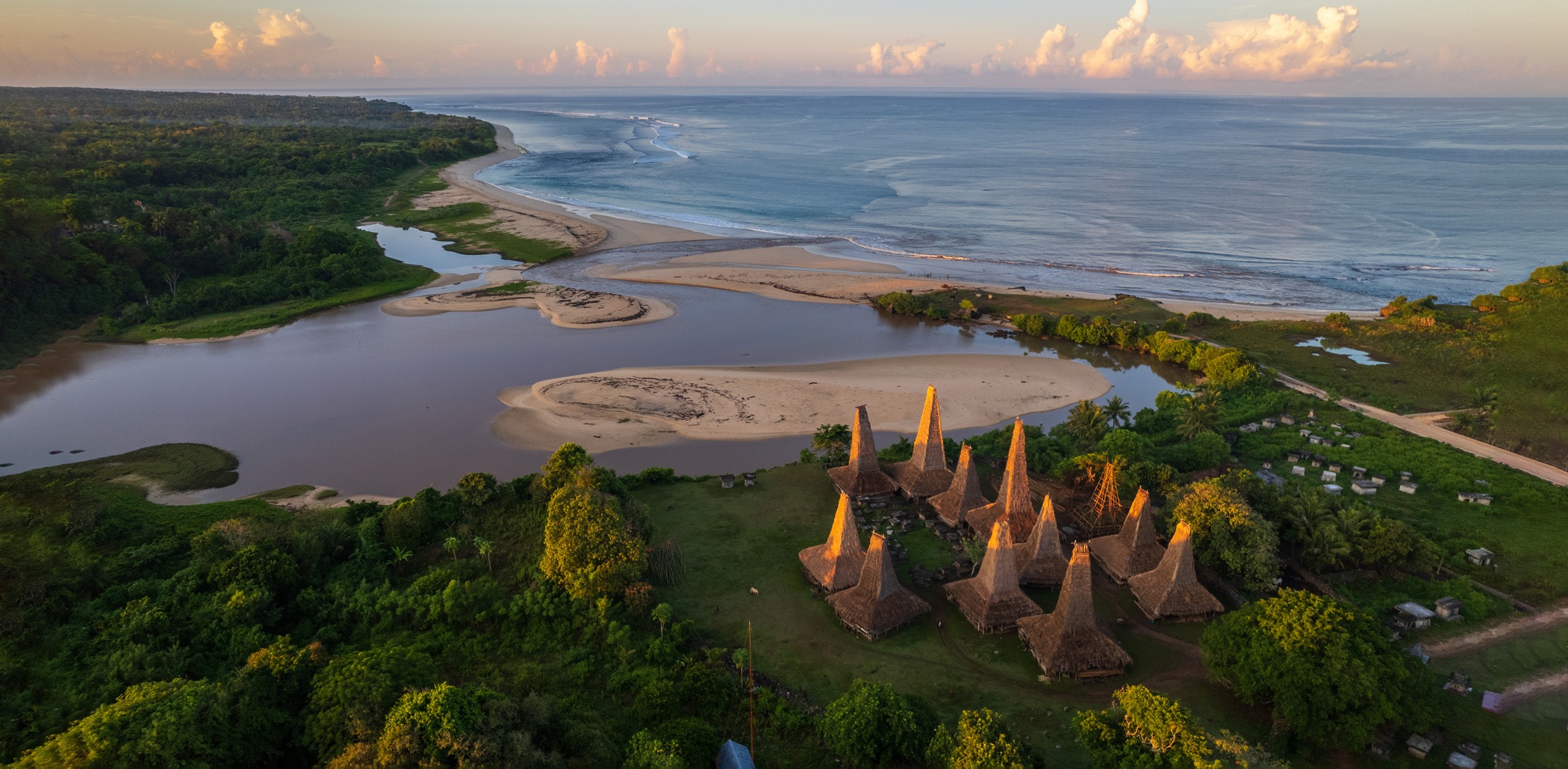 An aerial view shows a cluster of tall thatched houses beside an estuary and sandy coastline on Sumba, Indonesia.
