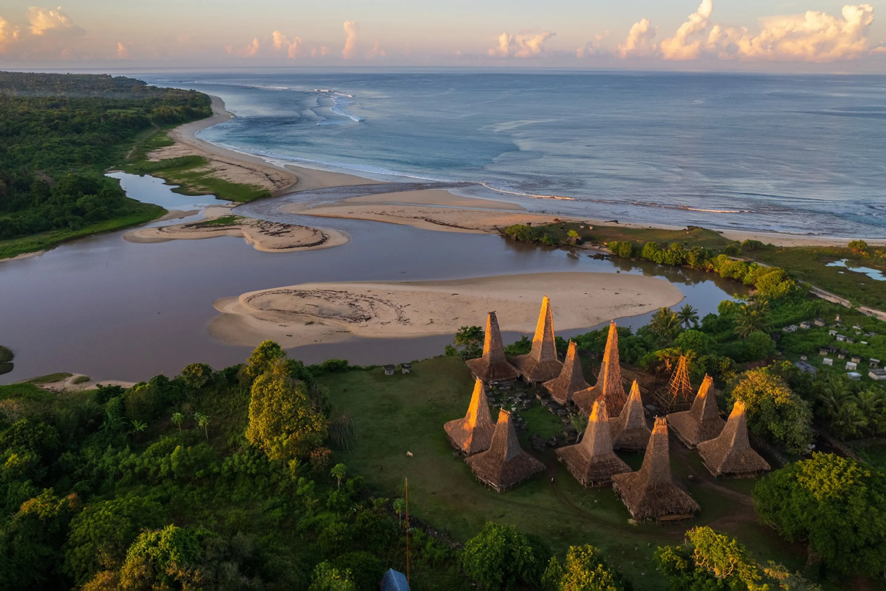 An aerial view shows a cluster of tall thatched houses beside an estuary and sandy coastline on Sumba, Indonesia.