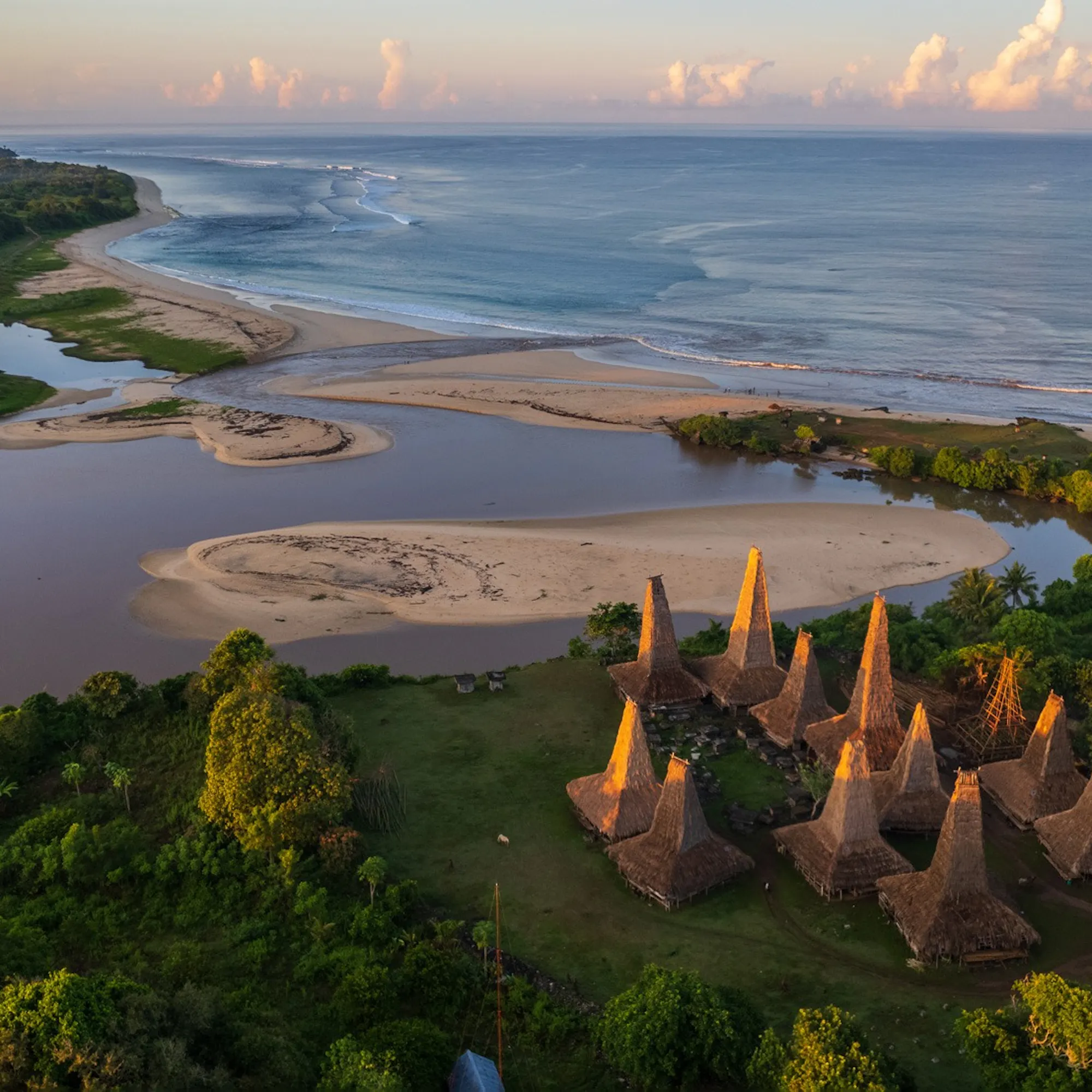 An aerial view shows a cluster of tall thatched houses beside an estuary and sandy coastline on Sumba, Indonesia.