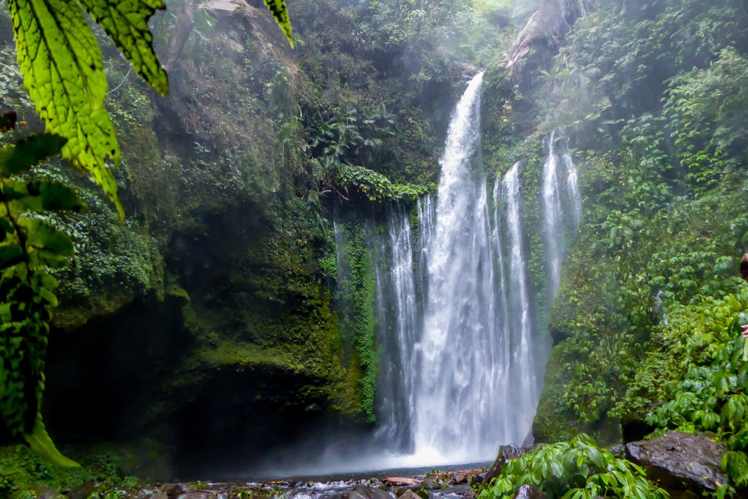 A tall waterfall drops into a rocky pool in Lombok, surrounded by dense green forest and dark wet stone walls.
