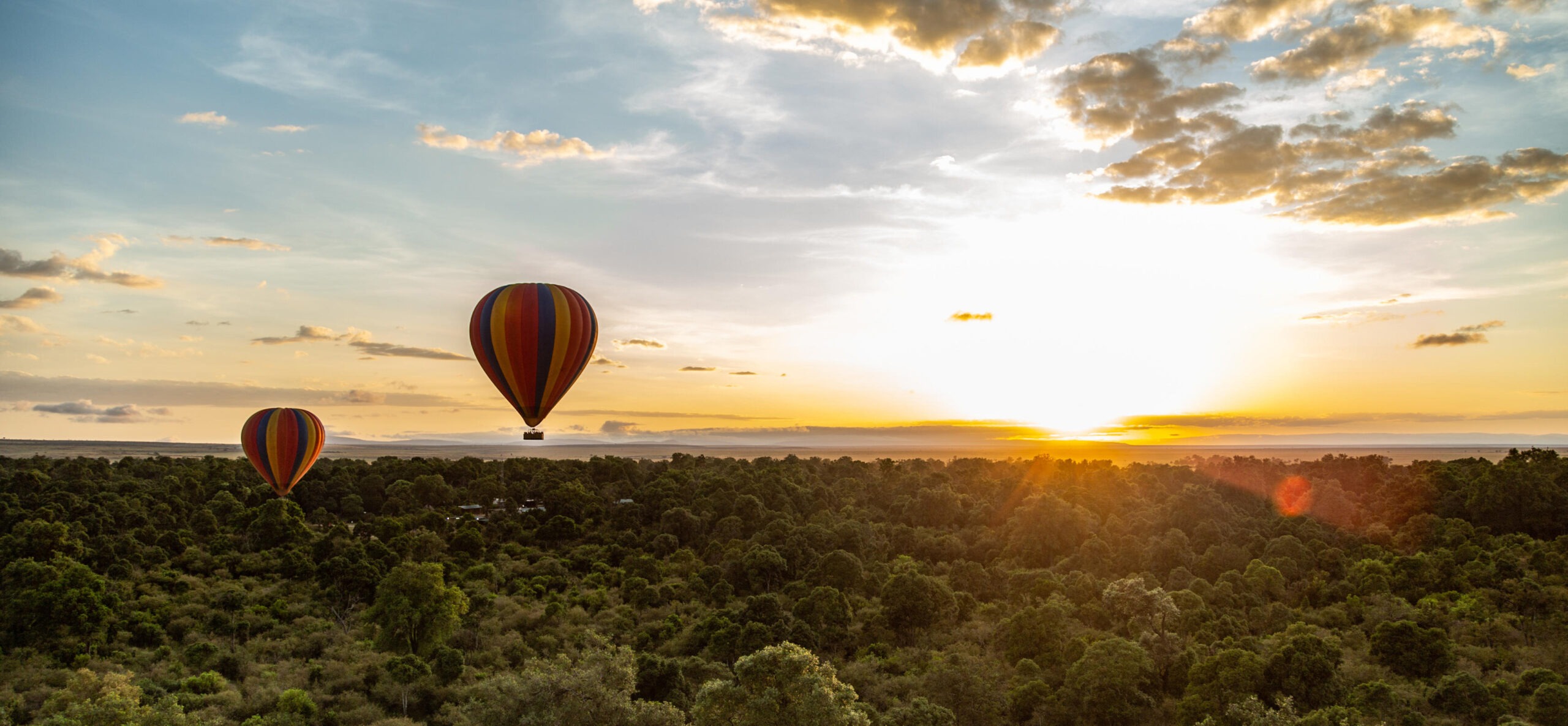 Two striped balloons float above dense treetops at sunrise, with low clouds and golden light over the Maasai Mara.