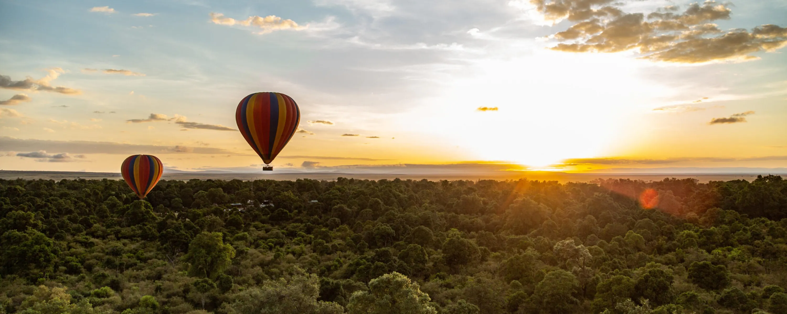 Two striped balloons float above dense treetops at sunrise, with low clouds and golden light over the Maasai Mara.