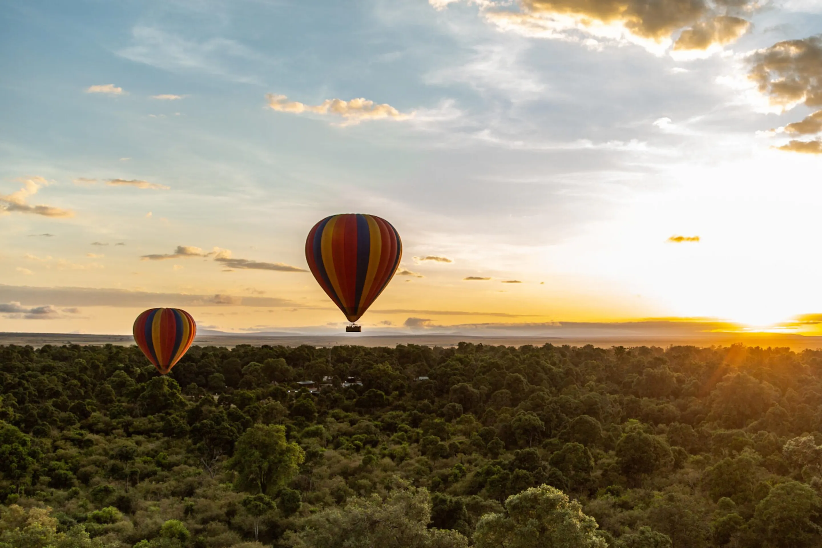 Two striped balloons float above dense treetops at sunrise, with low clouds and golden light over the Maasai Mara.