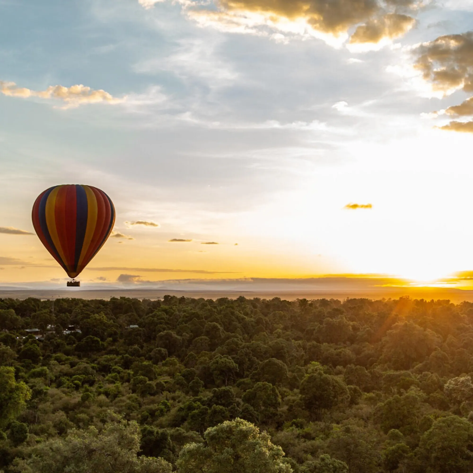 Two striped balloons float above dense treetops at sunrise, with low clouds and golden light over the Maasai Mara.