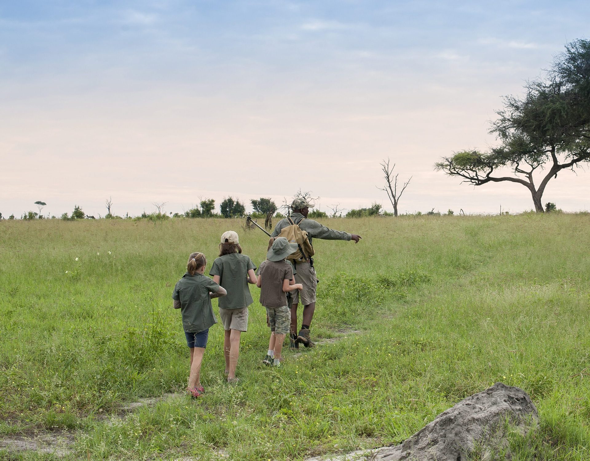 A walking guide leads three children along a grassy path, pointing across open plains beneath a wide pale sky.