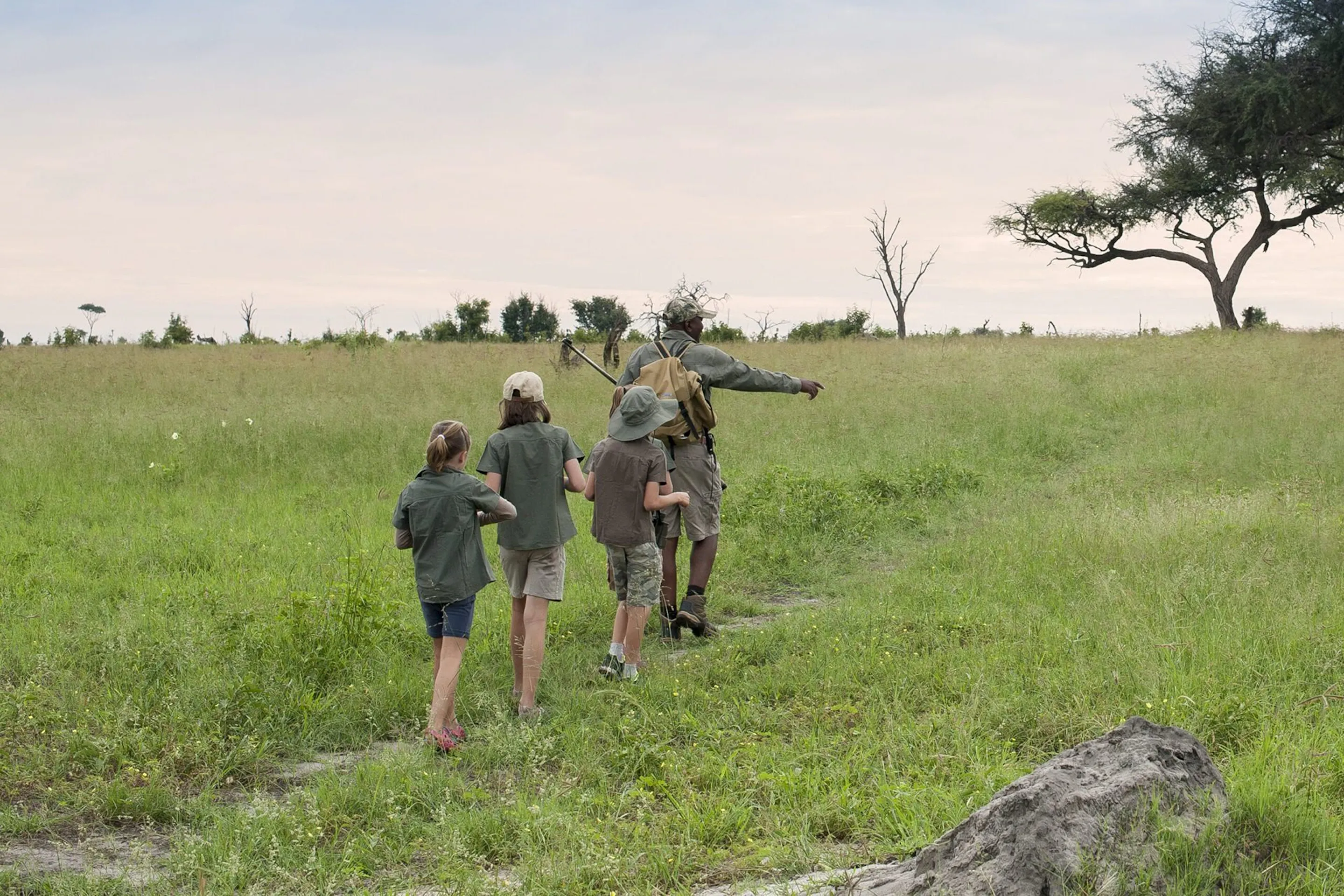 A walking guide leads three children along a grassy path, pointing across open plains beneath a wide pale sky.