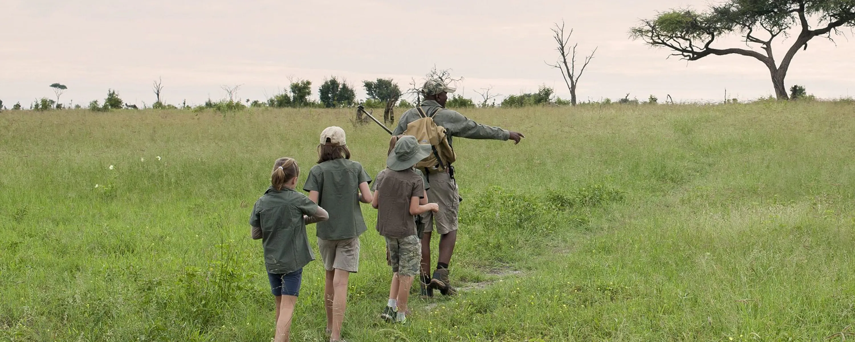 A walking guide leads three children along a grassy path, pointing across open plains beneath a wide pale sky.