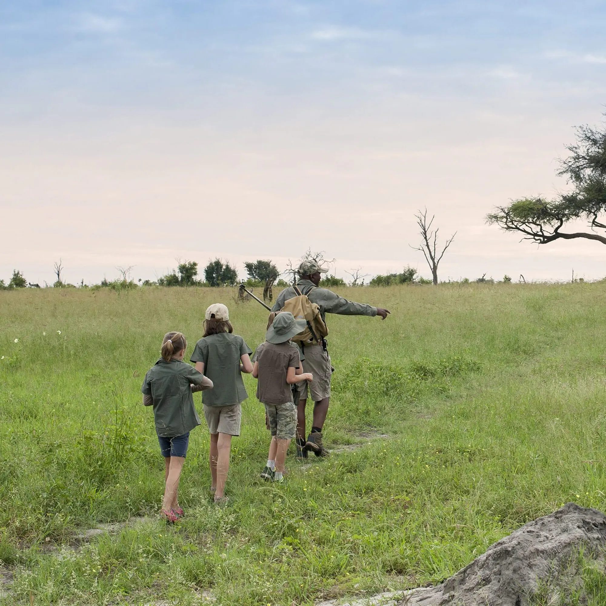 A walking guide leads three children along a grassy path, pointing across open plains beneath a wide pale sky.