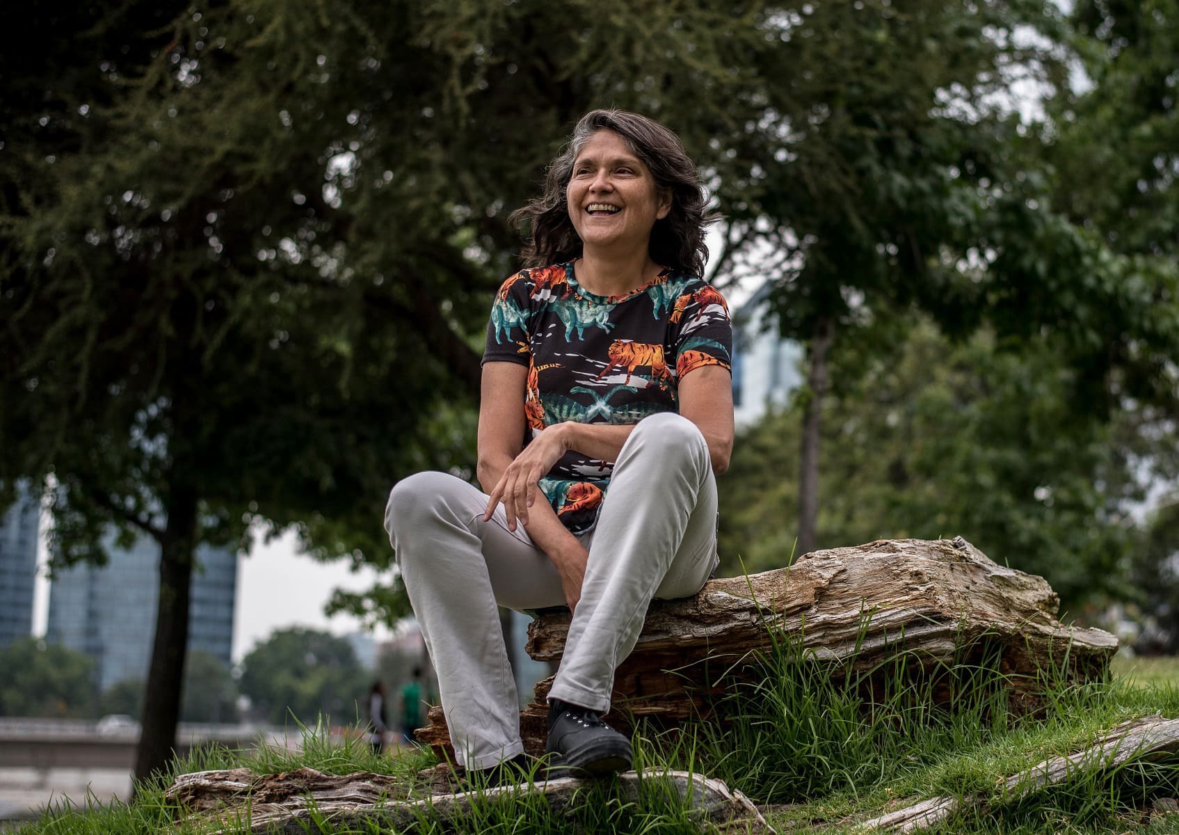 Bárbara Saavedra sits on a weathered log in a grassy park, with tall trees and blurred city buildings behind her.