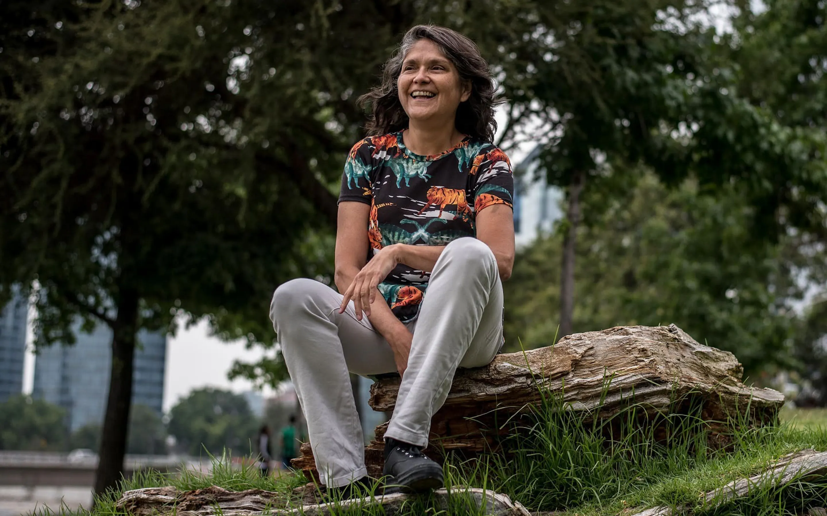 Bárbara Saavedra sits on a weathered log in a grassy park, with tall trees and blurred city buildings behind her.