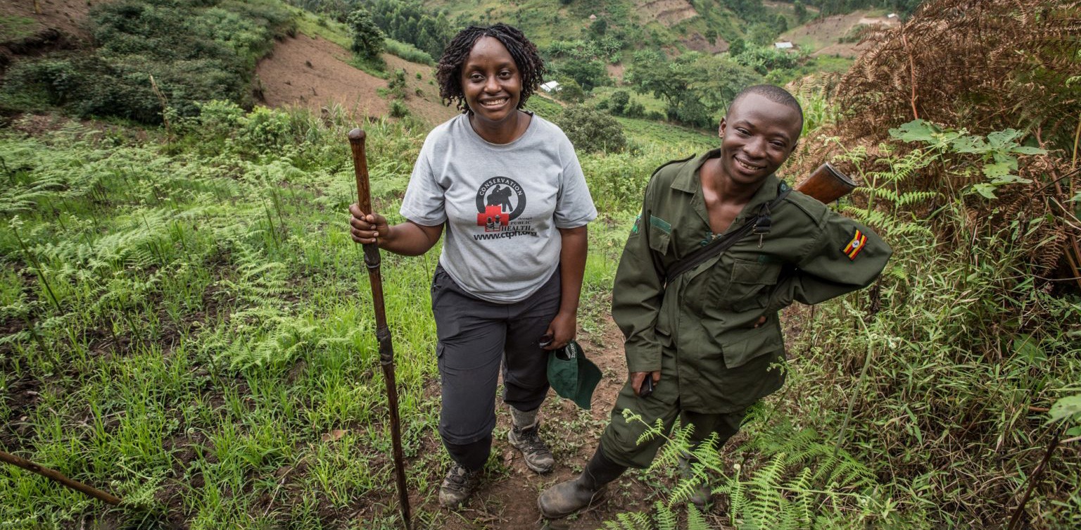 Dr. Gladys Kalema-Zikusoka stands on a steep green hillside trail beside a ranger in Uganda's forested highlands.