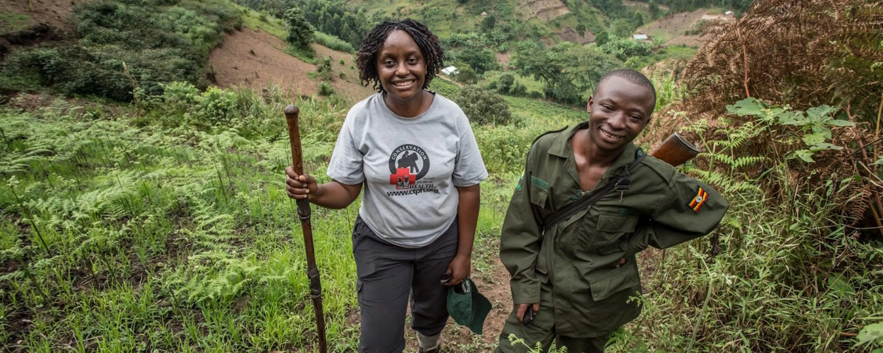 Dr. Gladys Kalema-Zikusoka stands on a steep green hillside trail beside a ranger in Uganda's forested highlands.