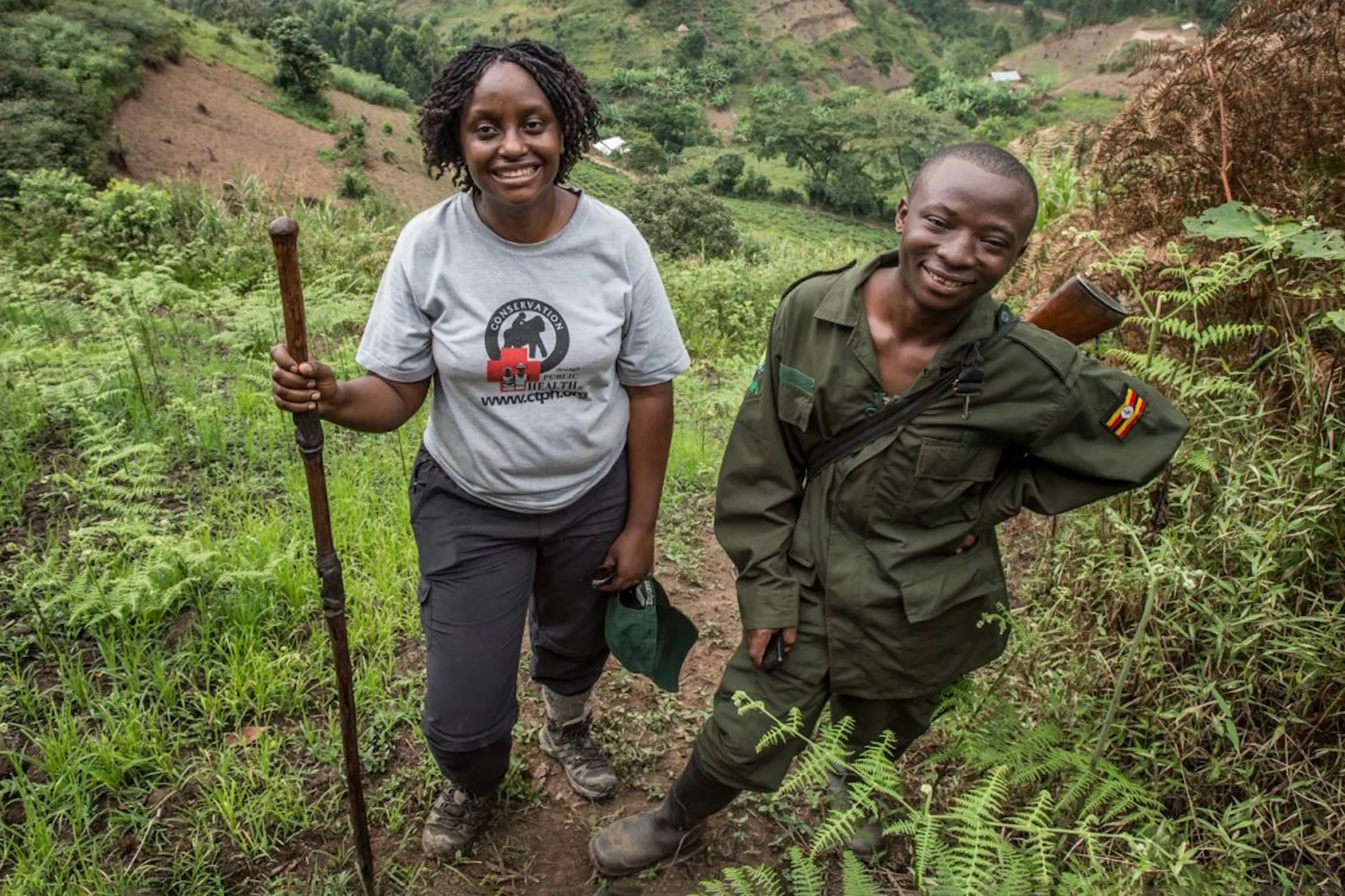 Dr. Gladys Kalema-Zikusoka stands on a steep green hillside trail beside a ranger in Uganda's forested highlands.