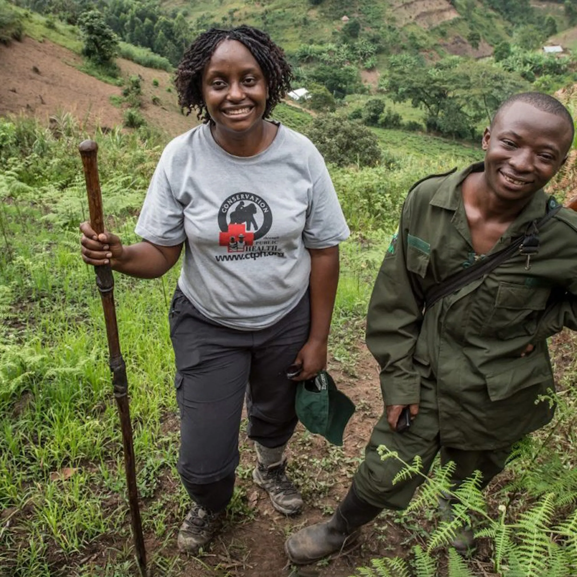 Dr. Gladys Kalema-Zikusoka stands on a steep green hillside trail beside a ranger in Uganda's forested highlands.