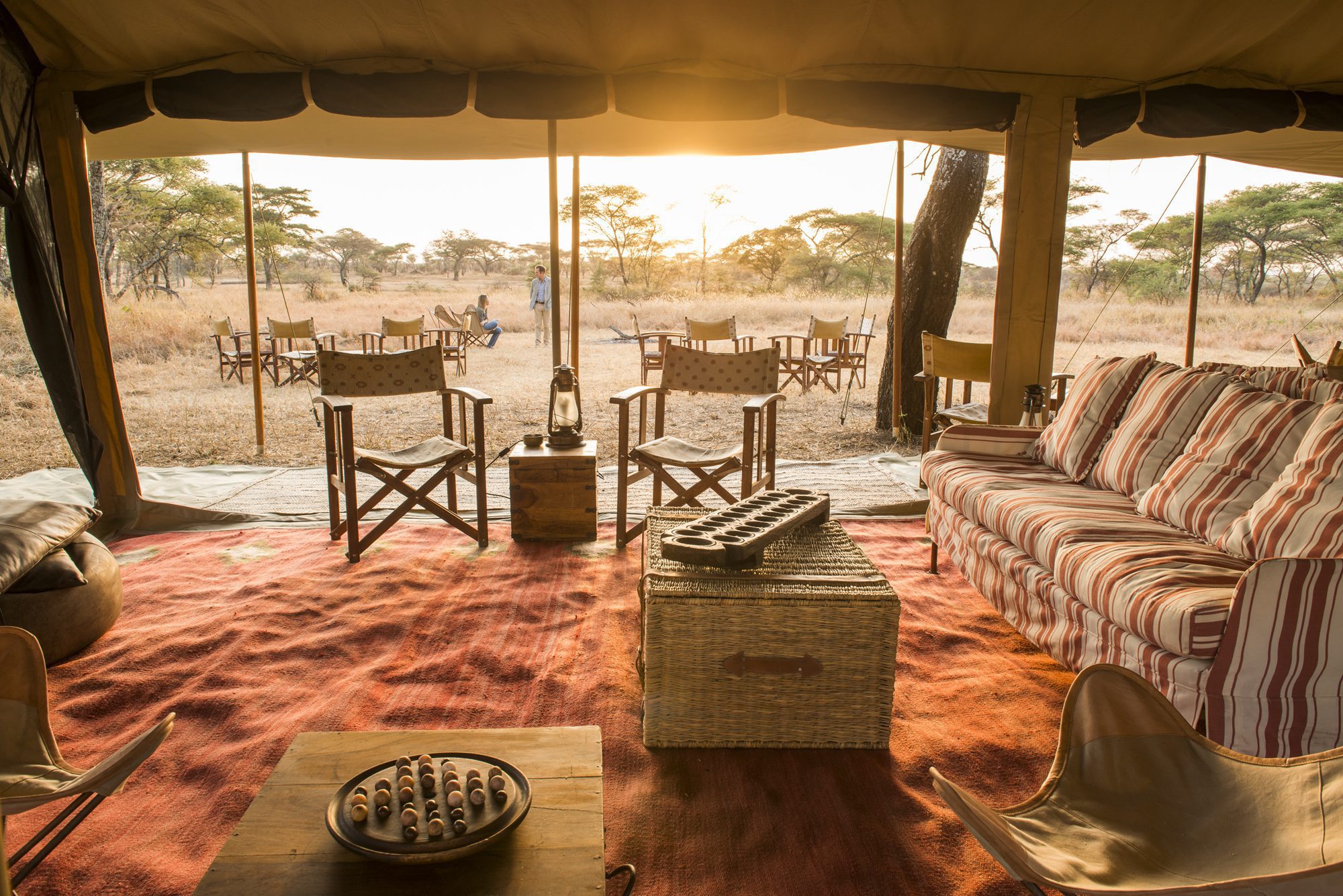 A tented lounge opens onto outdoor chairs and a set table, with warm sunrise light falling across the grassland.