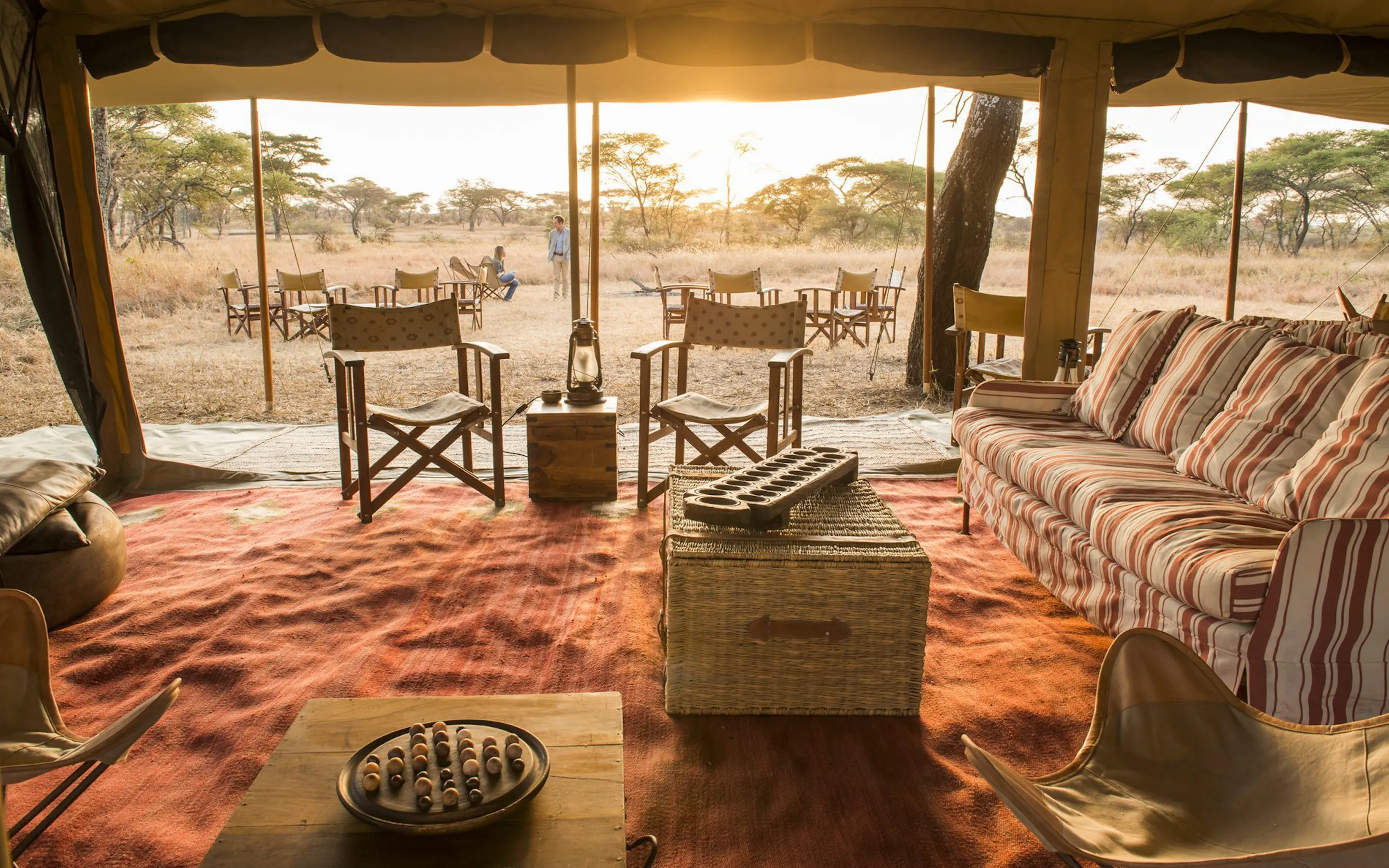 A tented lounge opens onto outdoor chairs and a set table, with warm sunrise light falling across the grassland.