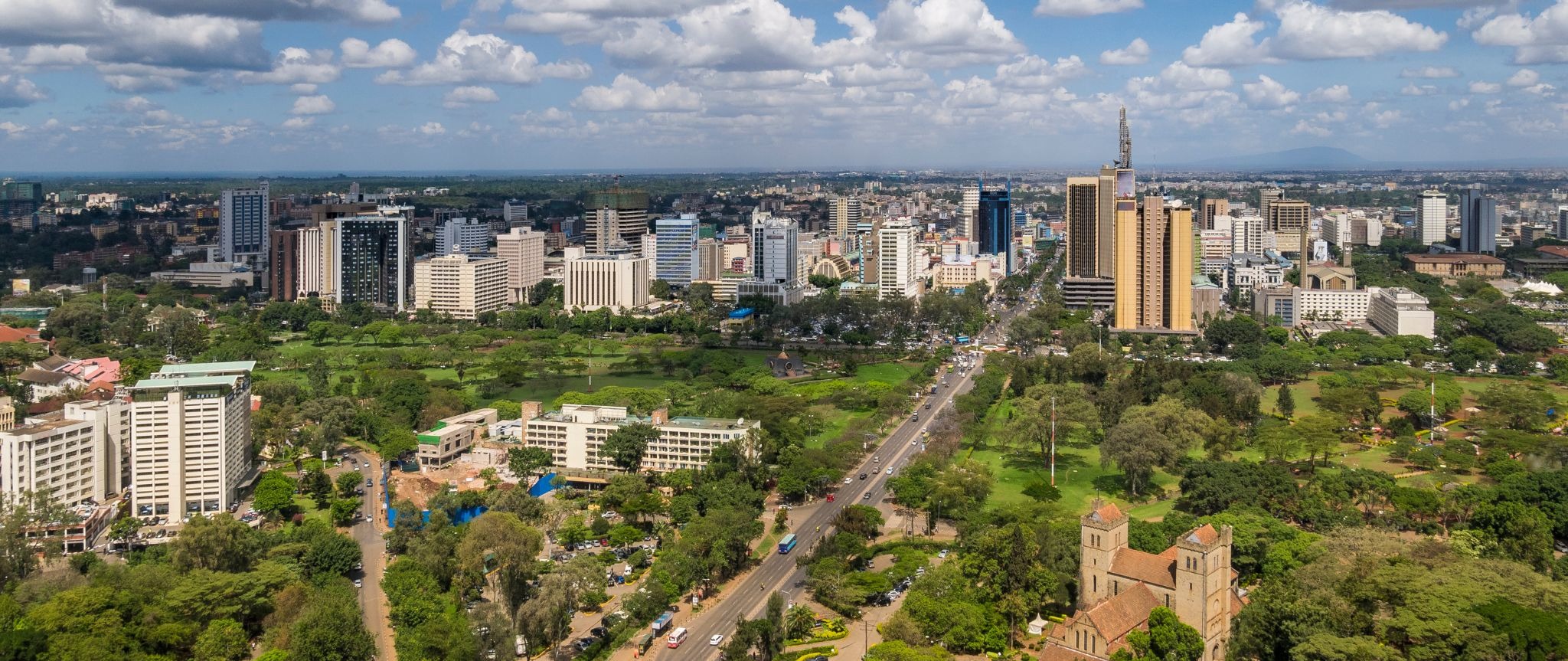 Nairobi's skyline stretches beyond a wide avenue, green parks, and a church beneath scattered clouds in Kenya.