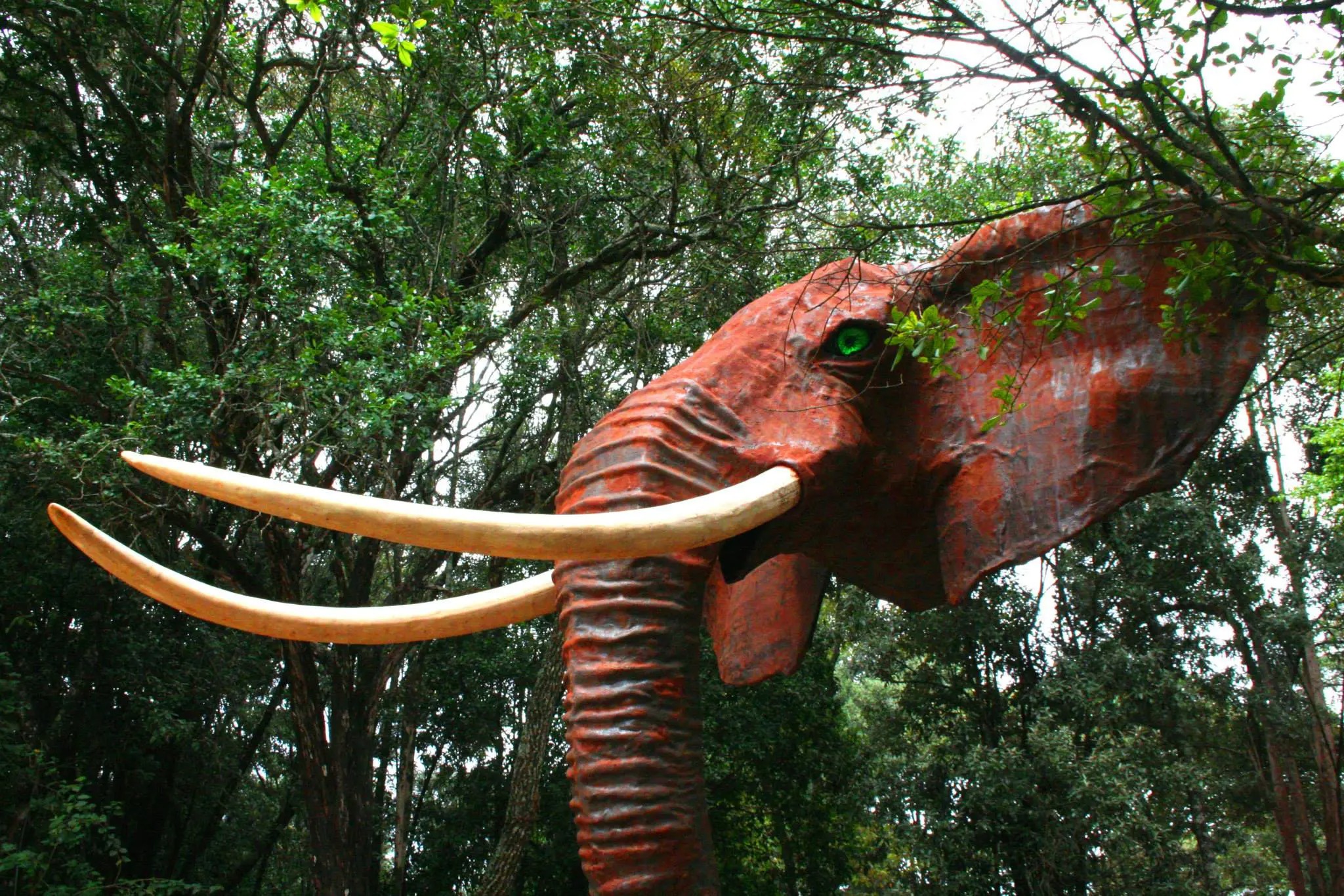 A red elephant sculpture with long pale tusks rises among dense trees at Matbronze Wildlife Art in Nairobi, Kenya.