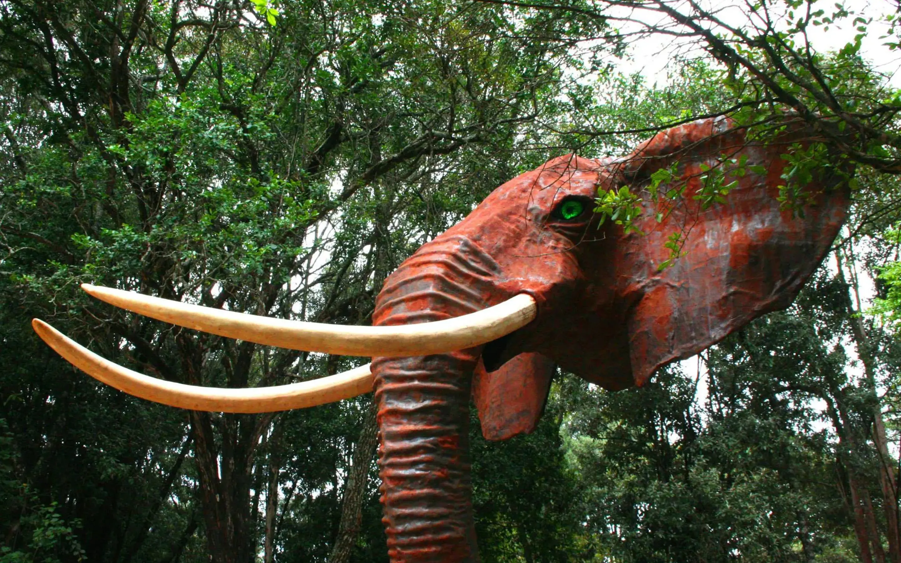 A red elephant sculpture with long pale tusks rises among dense trees at Matbronze Wildlife Art in Nairobi, Kenya.