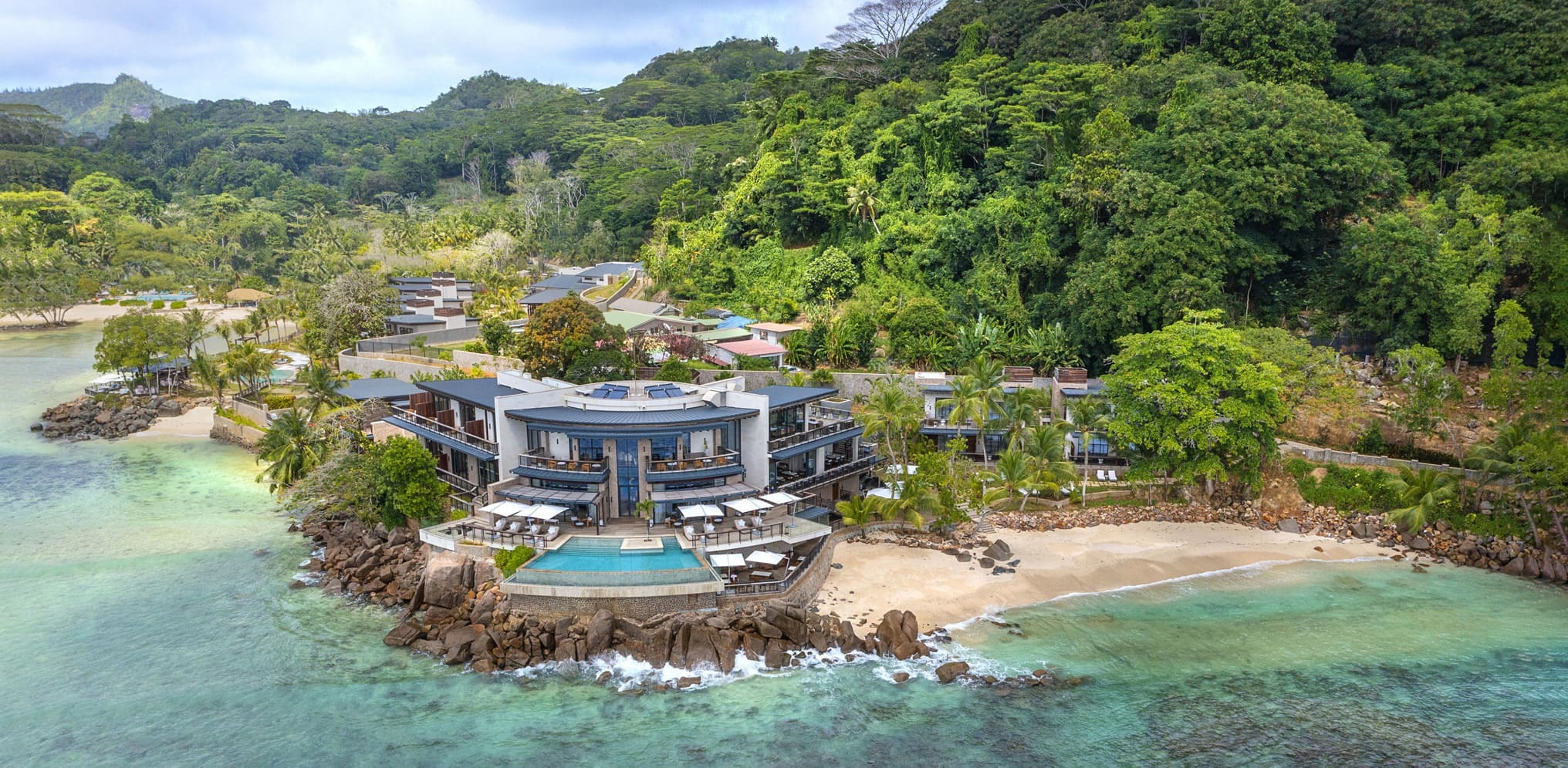 A coastal resort with pools and villas sits beside turquoise water and forested hills on Mahé in the Seychelles.