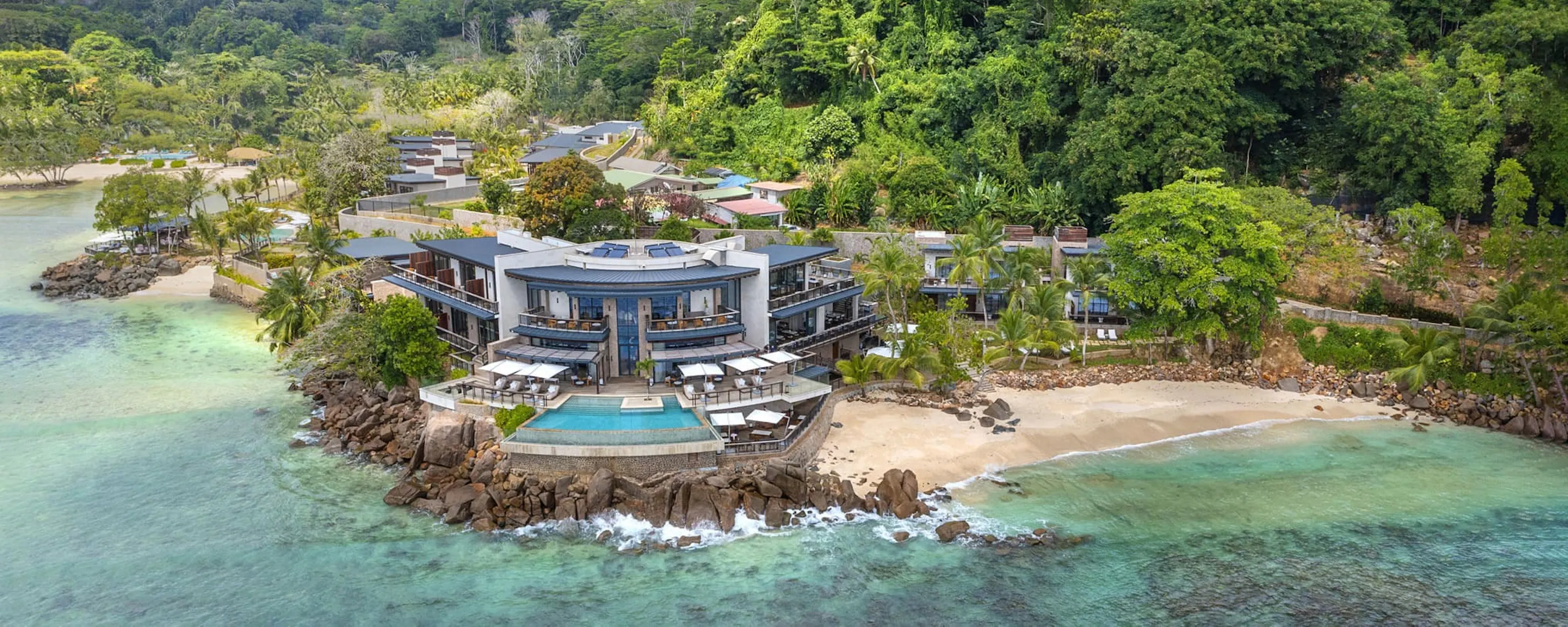 A coastal resort with pools and villas sits beside turquoise water and forested hills on Mahé in the Seychelles.