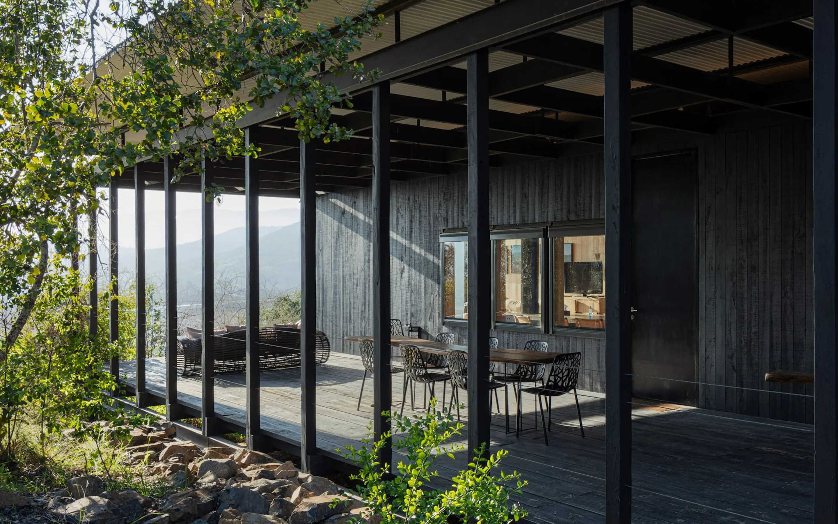 A shaded timber terrace with dining tables overlooks hazy mountain views at Vik Chile in the Cachapoal Valley.