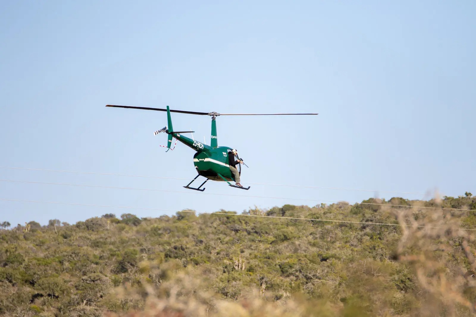 A green helicopter flies low above scrubland during a rhino darting and monitoring operation in South Africa.