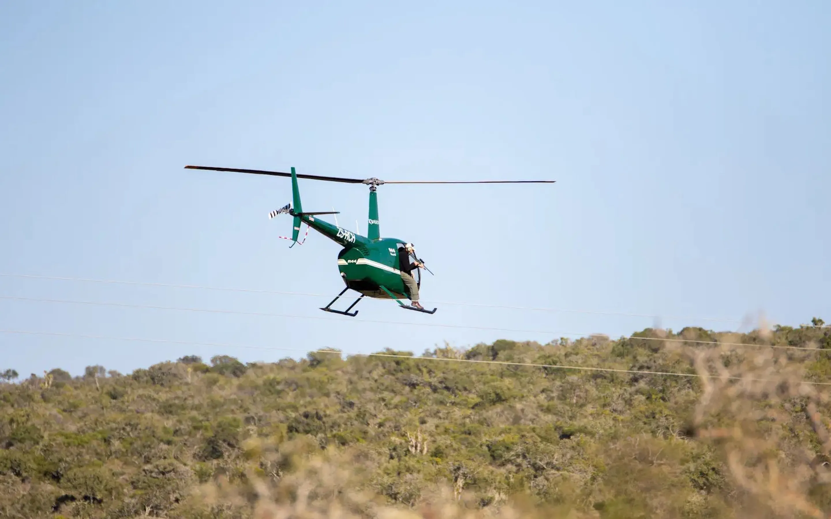 A green helicopter flies low above scrubland during a rhino darting and monitoring operation in South Africa.