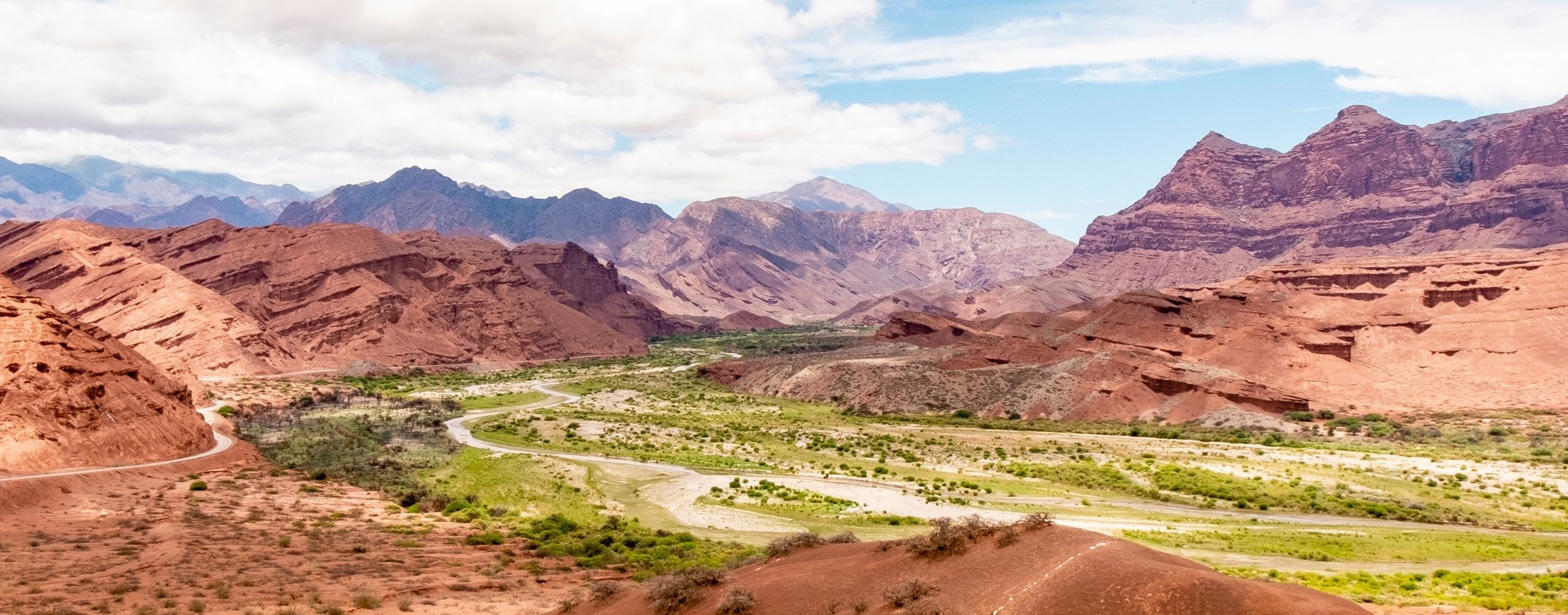 A winding road cuts through a green valley between red rock hills and jagged mountains near Cafayate in Argentina.