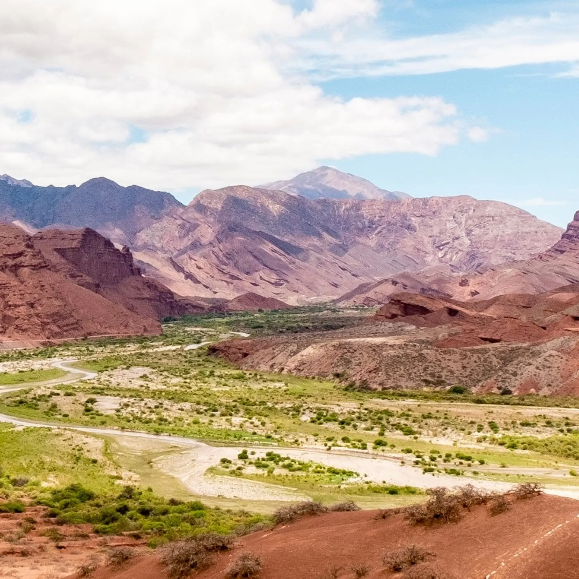 A winding road cuts through a green valley between red rock hills and jagged mountains near Cafayate in Argentina.