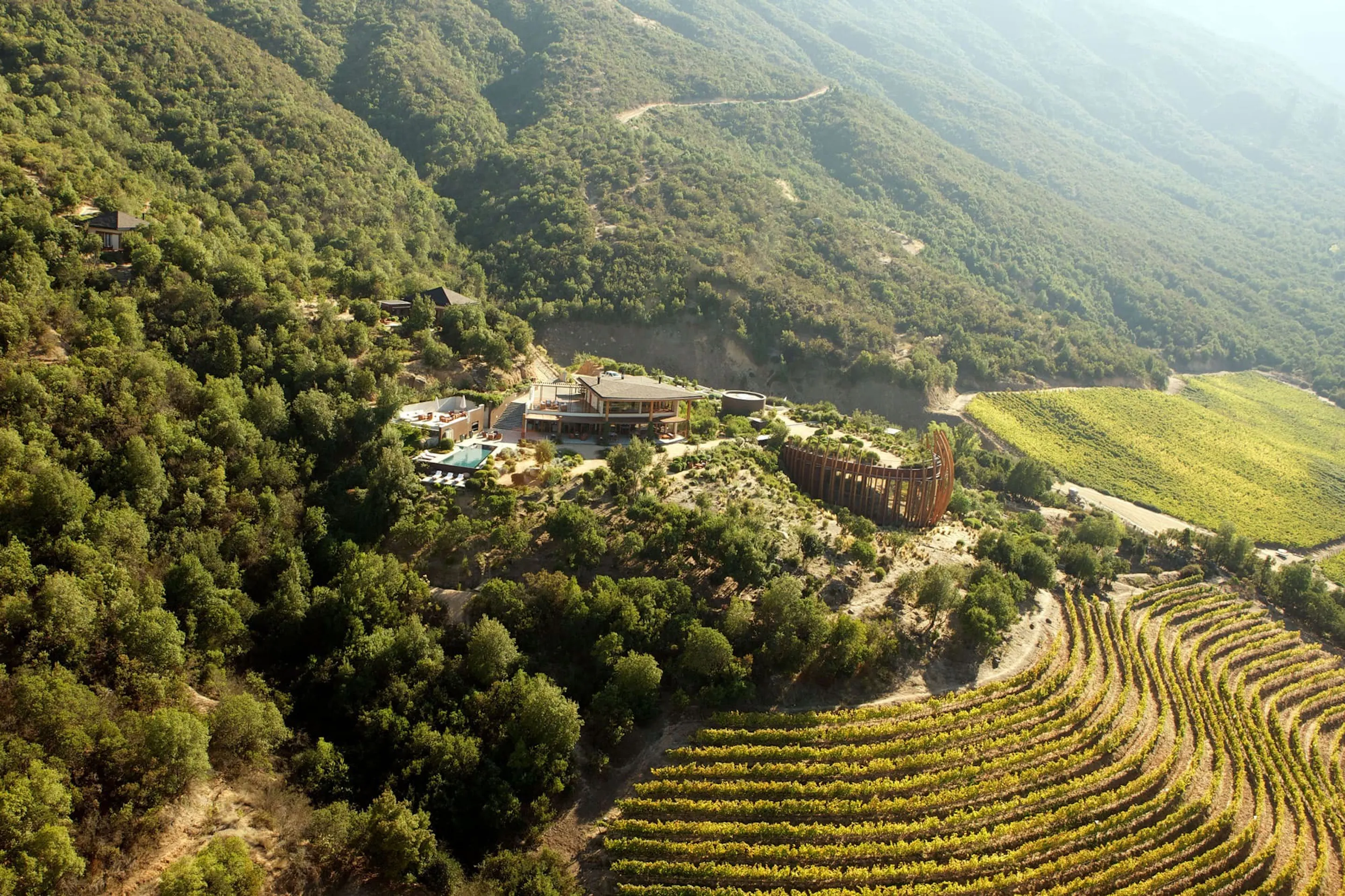 An aerial view shows Clos Apalta residence and vineyard rows tucked into forested hills in Chile's Colchagua Valley.