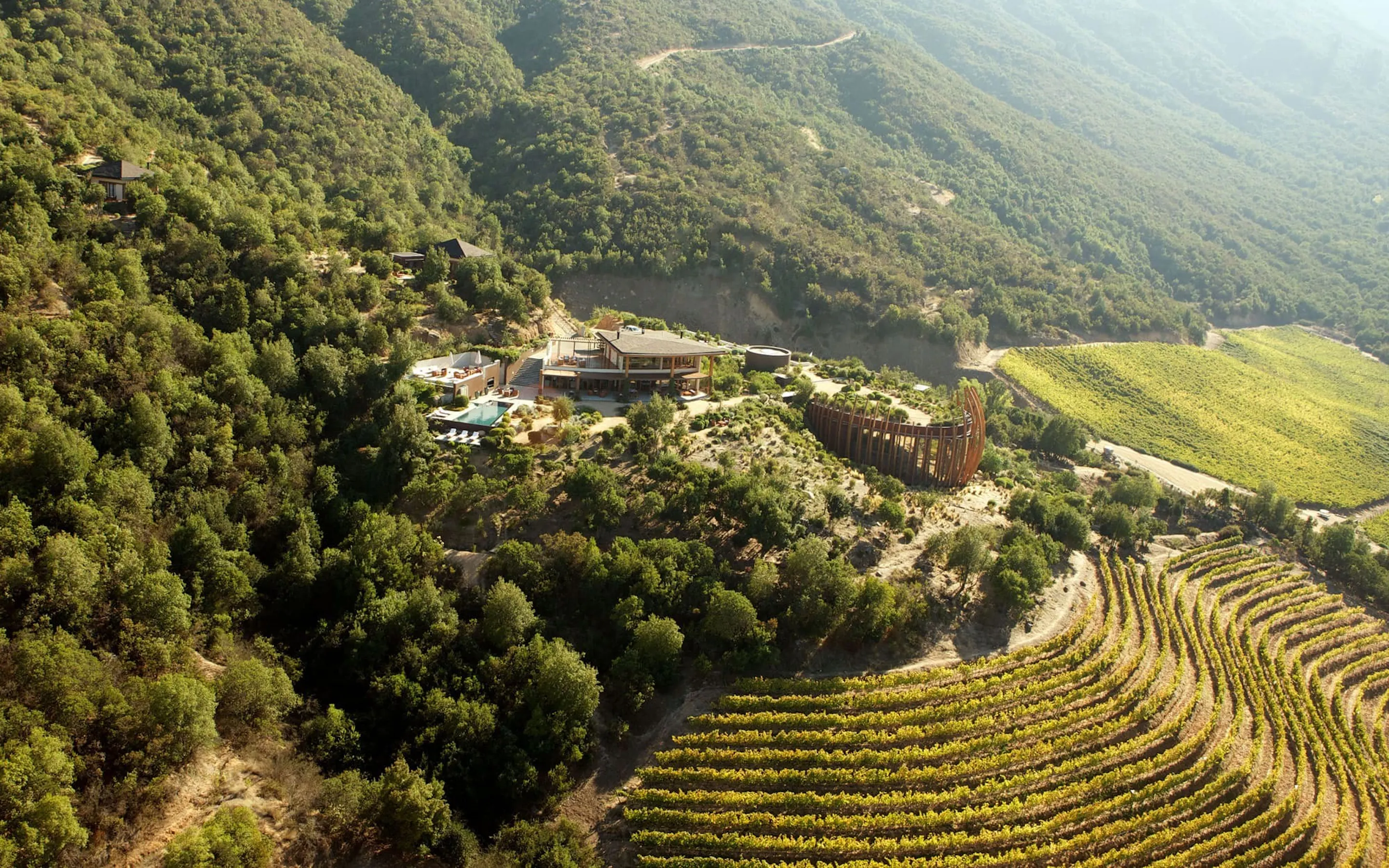 An aerial view shows Clos Apalta residence and vineyard rows tucked into forested hills in Chile's Colchagua Valley.