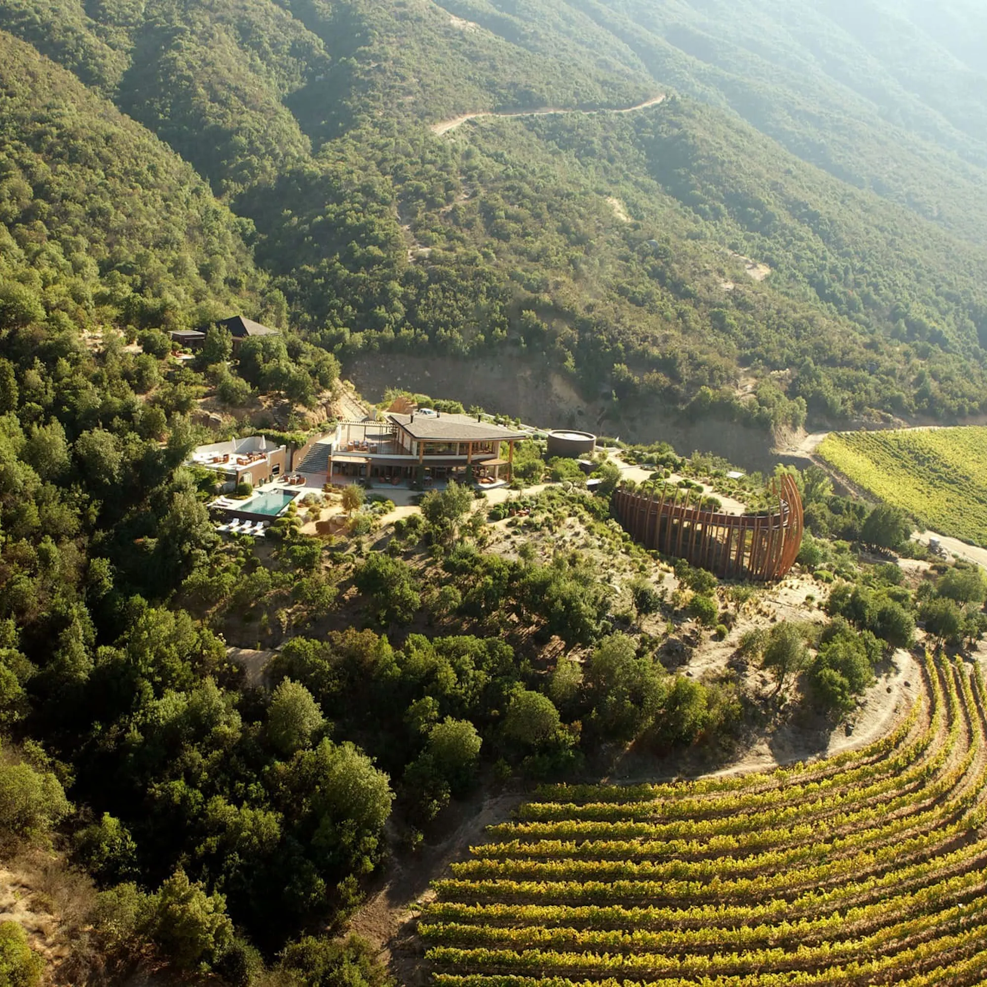 An aerial view shows Clos Apalta residence and vineyard rows tucked into forested hills in Chile's Colchagua Valley.