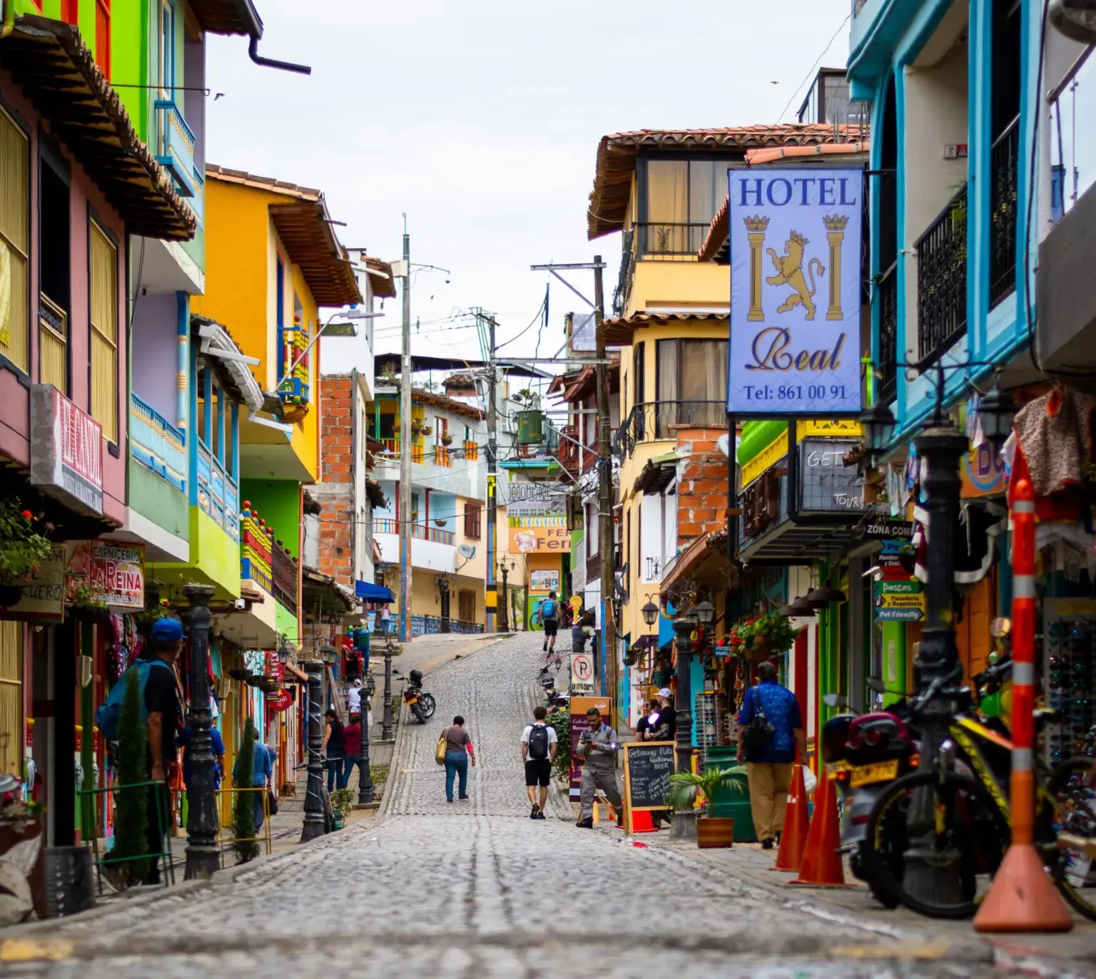 A cobbled street slopes past colorful buildings, hotel signs, parked motorbikes, and pedestrians in Guatapé, Colombia.