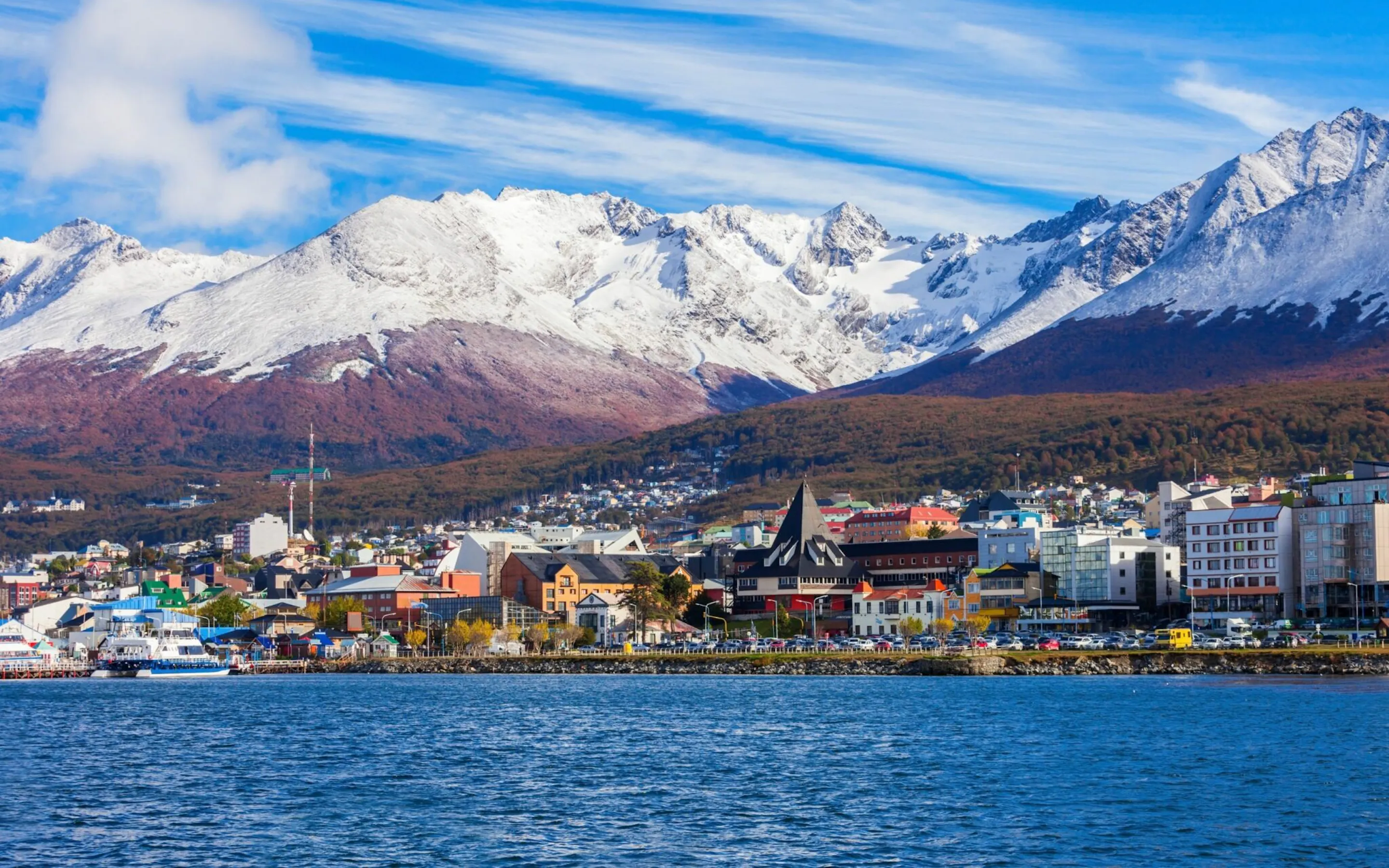 Ushuaia's waterfront stretches below snowy mountains, with colorful buildings lining the shore in Patagonia.
