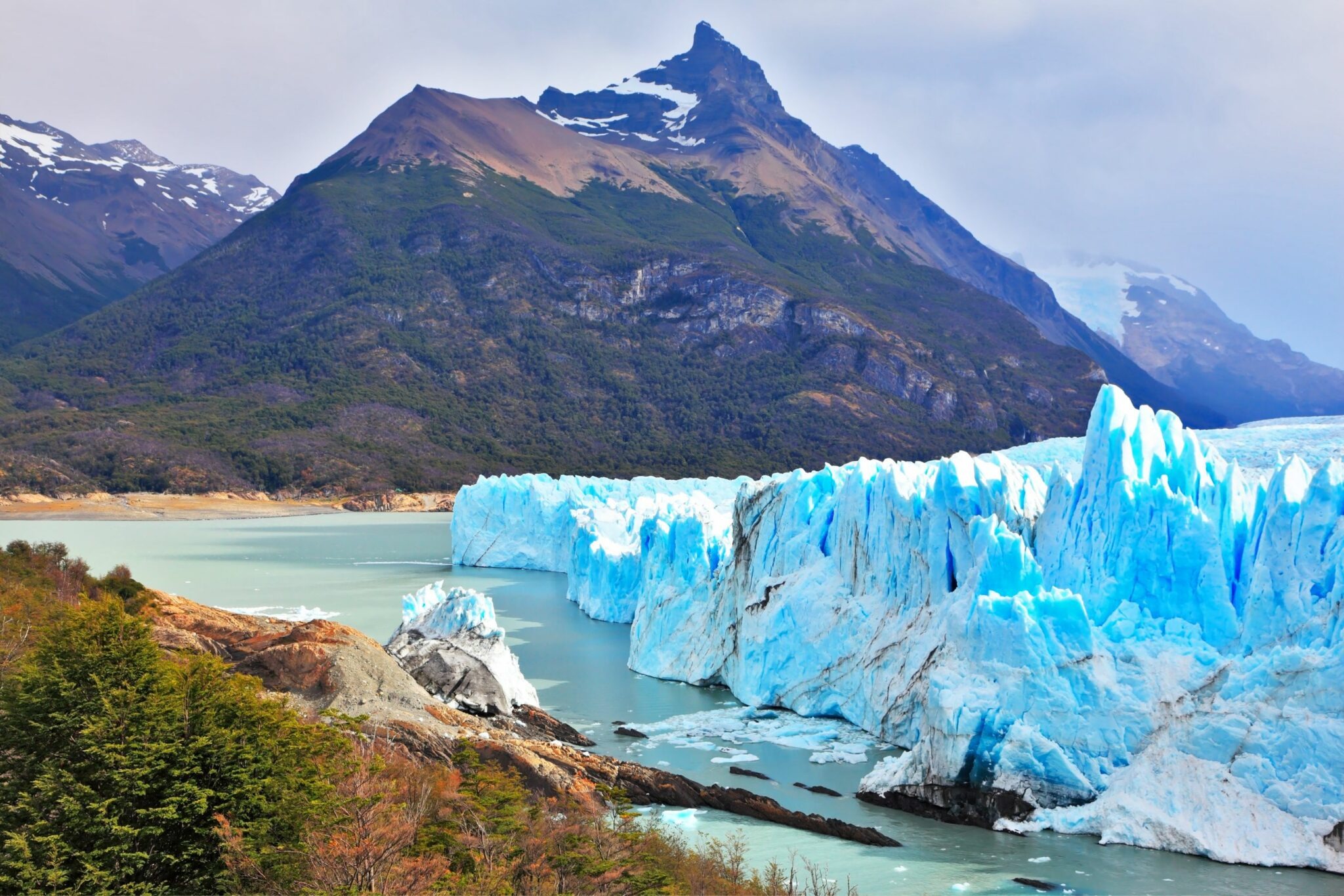 The blue face of Perito Moreno Glacier rises above a lake, framed by forest and dark peaks in Patagonia, Argentina.