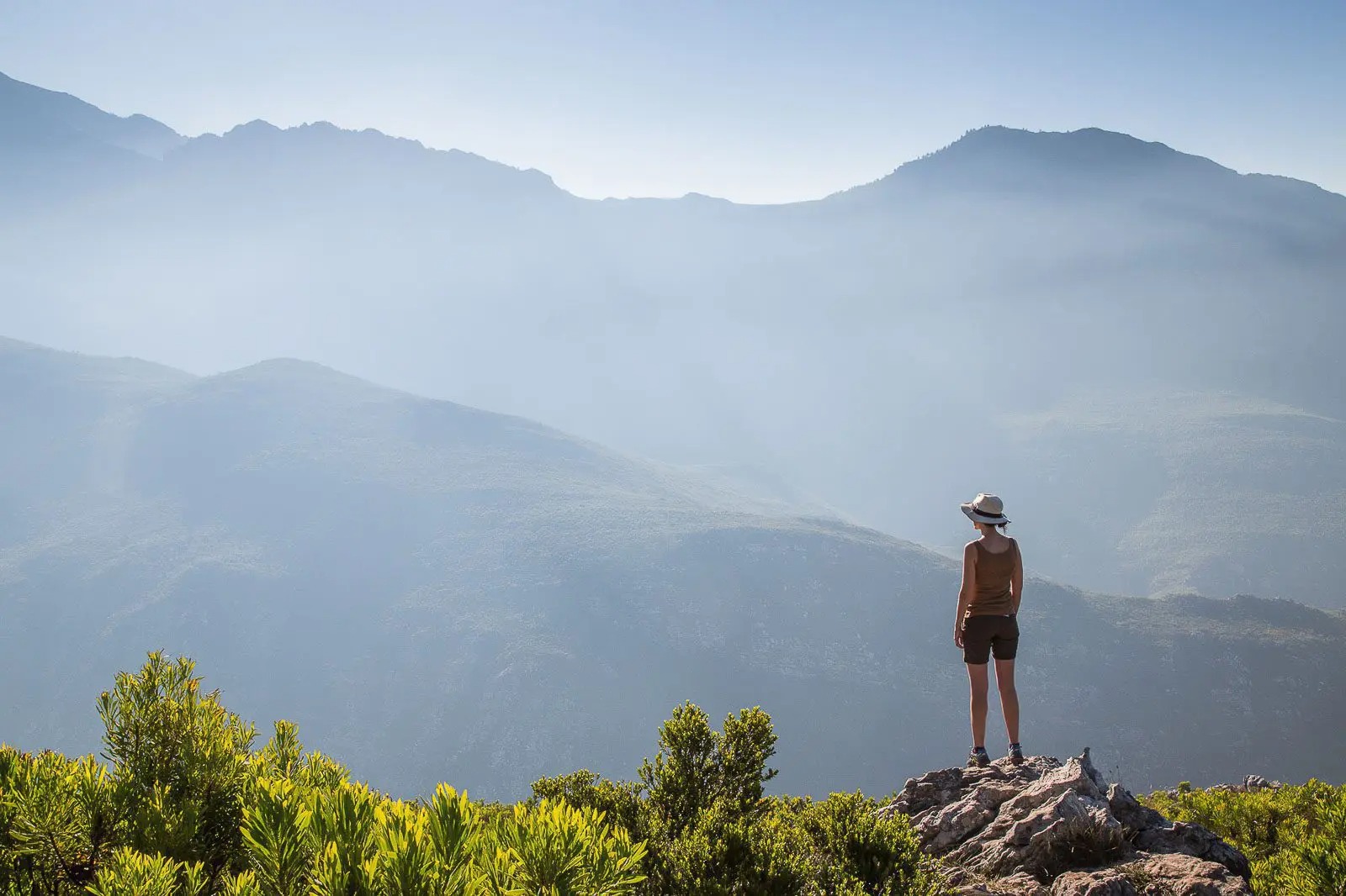 A woman in a brimmed hat stands on a rocky peak above misty mountain ranges in South Africa's Cape Winelands.