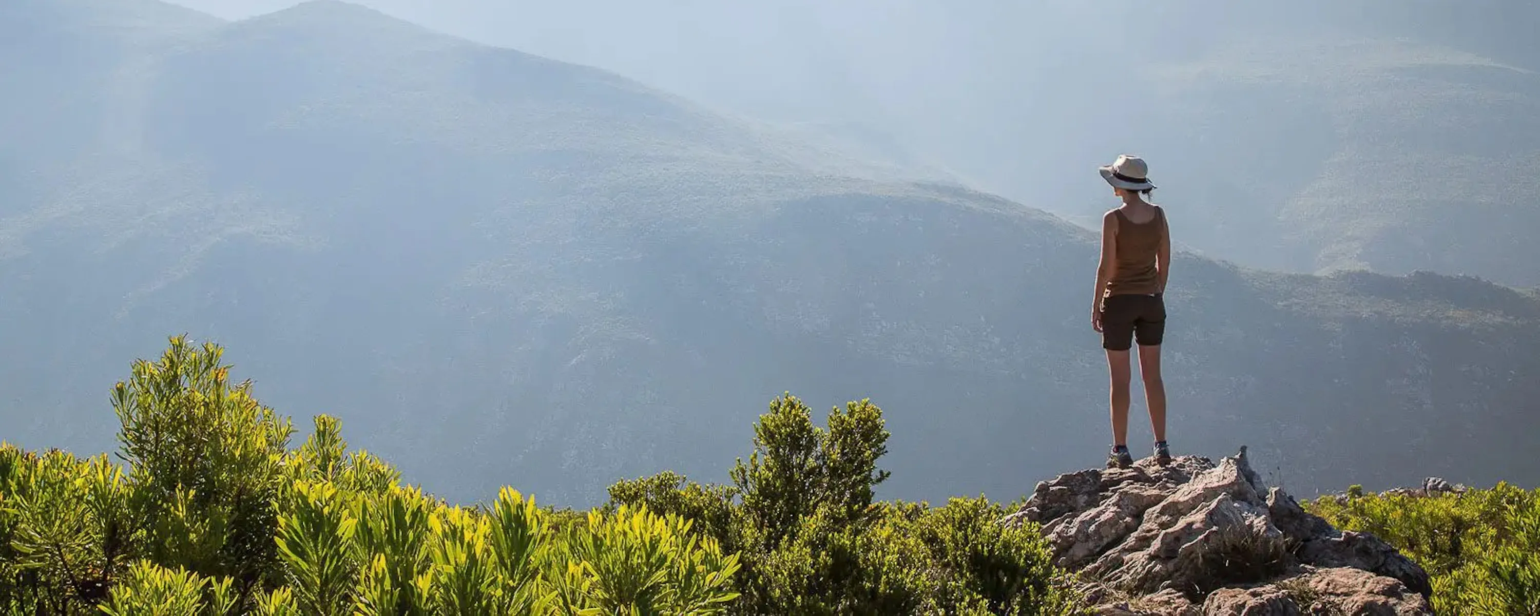 A woman in a brimmed hat stands on a rocky peak above misty mountain ranges in South Africa's Cape Winelands.