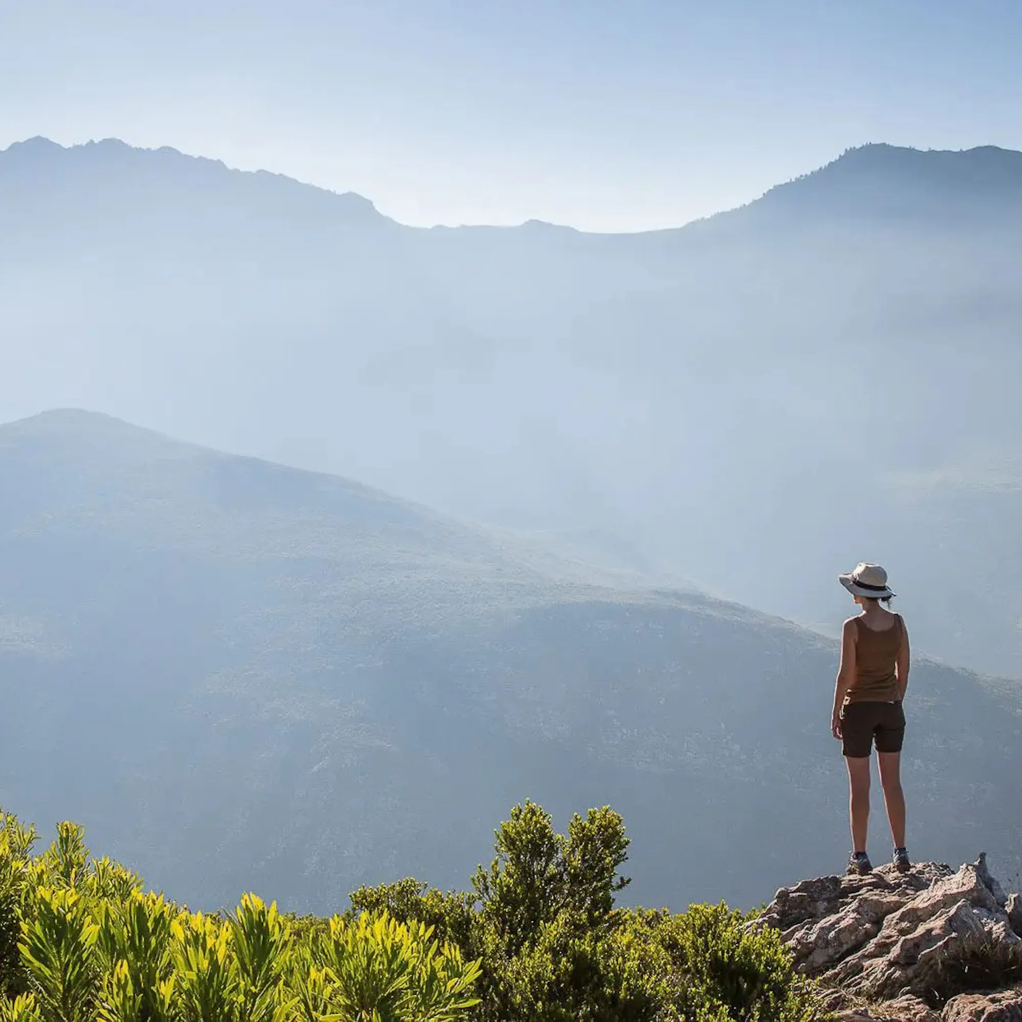 A woman in a brimmed hat stands on a rocky peak above misty mountain ranges in South Africa's Cape Winelands.