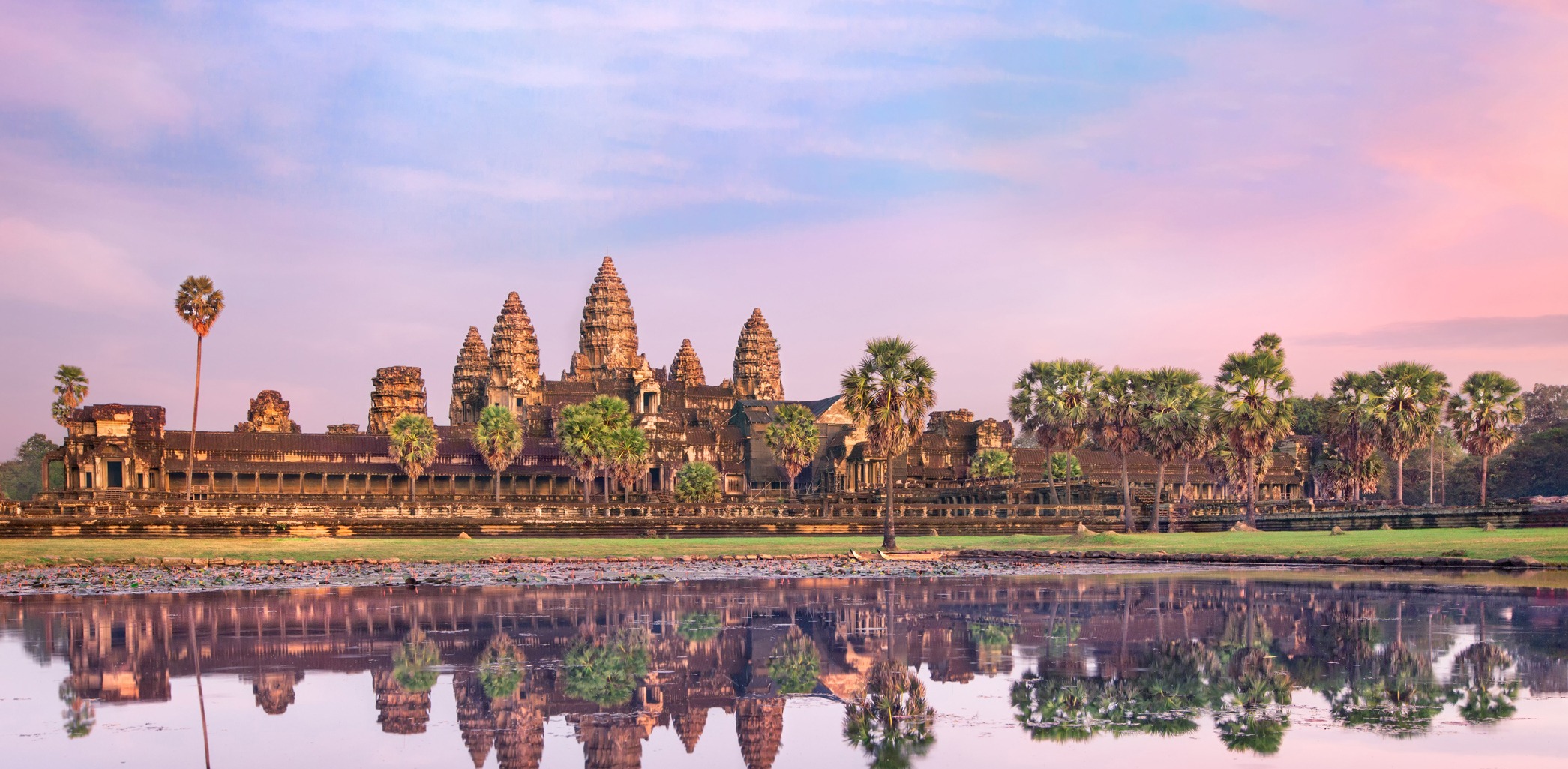 Weathered stone faces and a temple gate rise above lush trees at Angkor Thom in Cambodia beneath a pale sky.