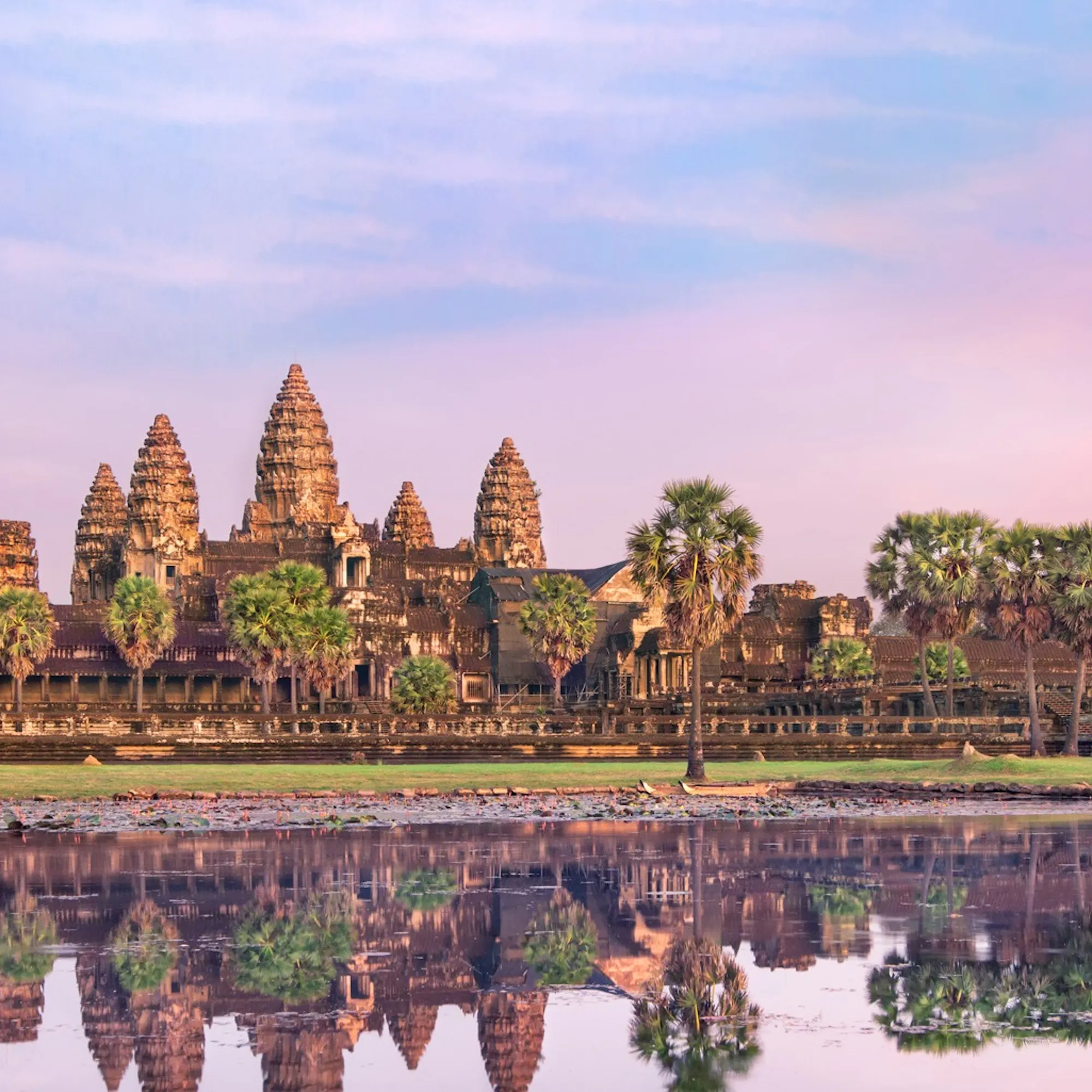 Weathered stone faces and a temple gate rise above lush trees at Angkor Thom in Cambodia beneath a pale sky.