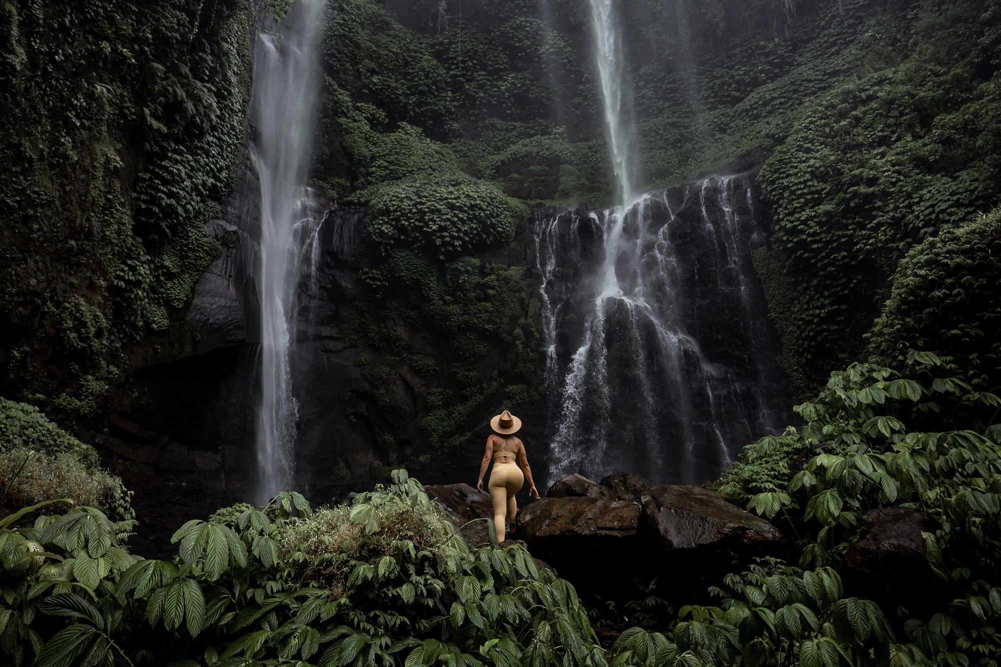 A traveler in a wide-brimmed hat stands on rocks before a tall, ivy-draped waterfall in Bali's lush jungle.