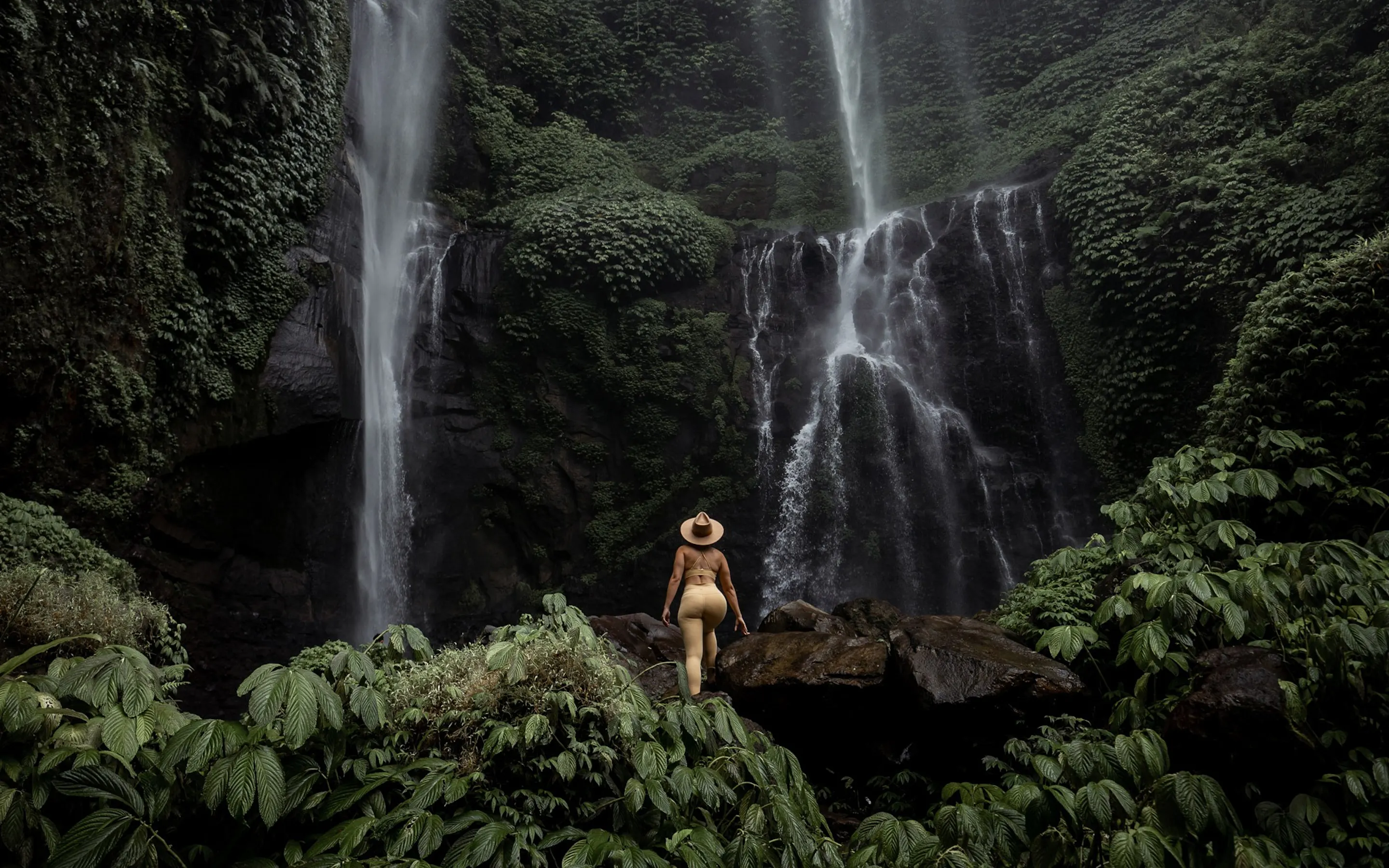 A traveler in a wide-brimmed hat stands on rocks before a tall, ivy-draped waterfall in Bali's lush jungle.