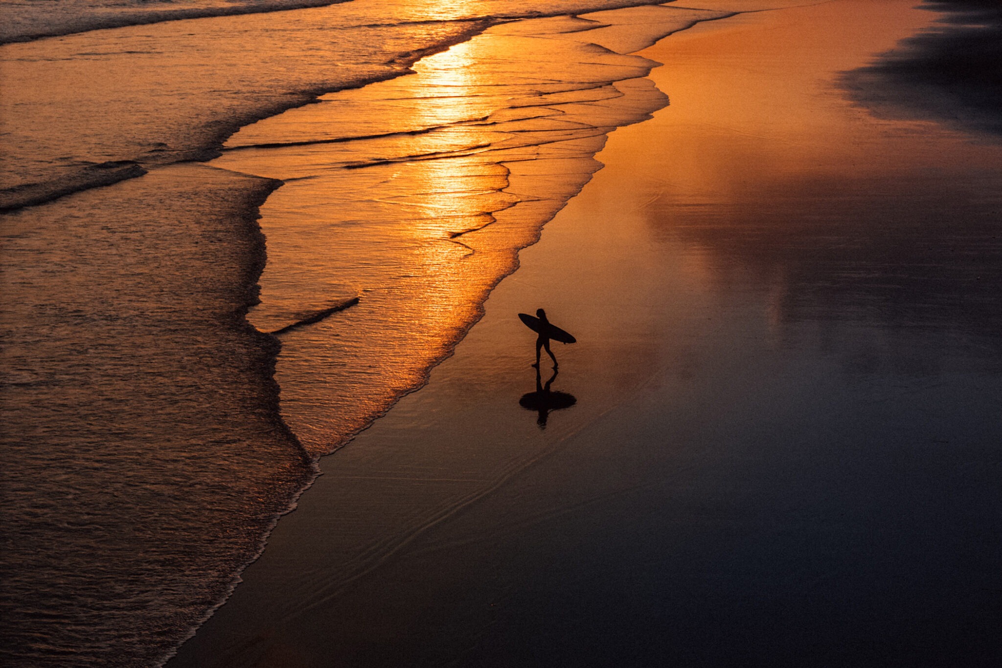 A lone surfer carries a board along wet sand as sunset light glows across the shoreline and receding waves in Bali.