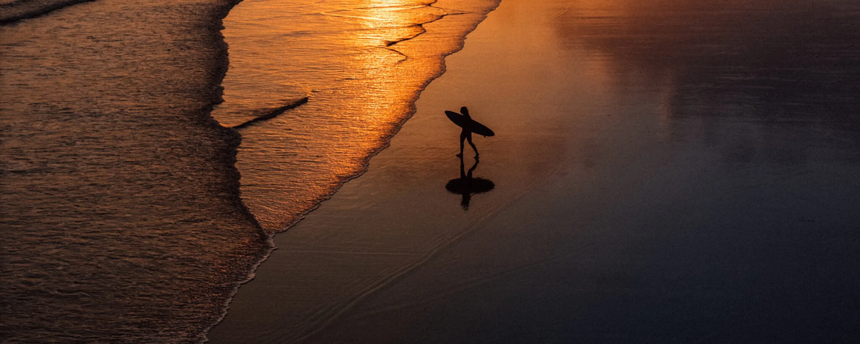 A lone surfer carries a board along wet sand as sunset light glows across the shoreline and receding waves in Bali.