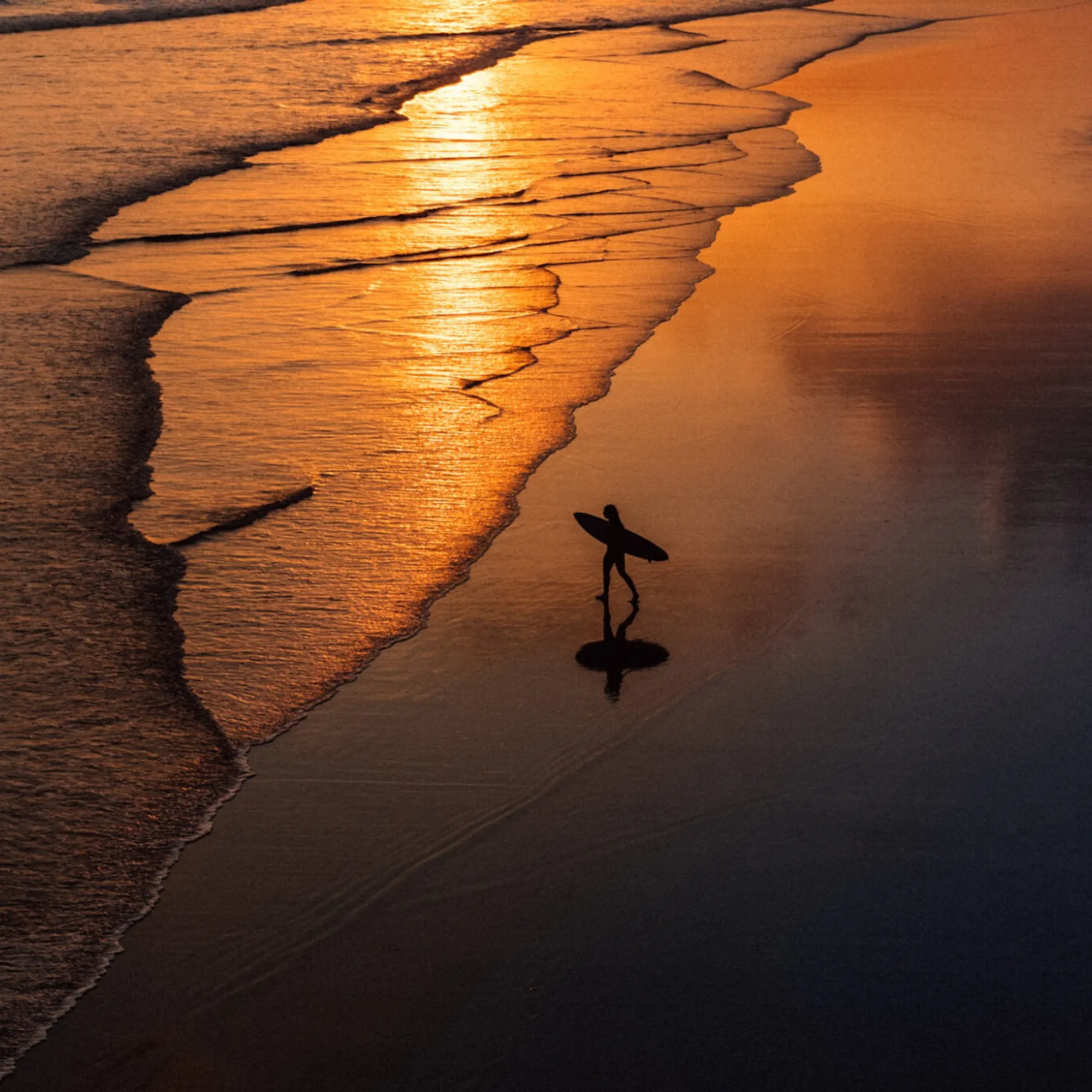 A lone surfer carries a board along wet sand as sunset light glows across the shoreline and receding waves in Bali.