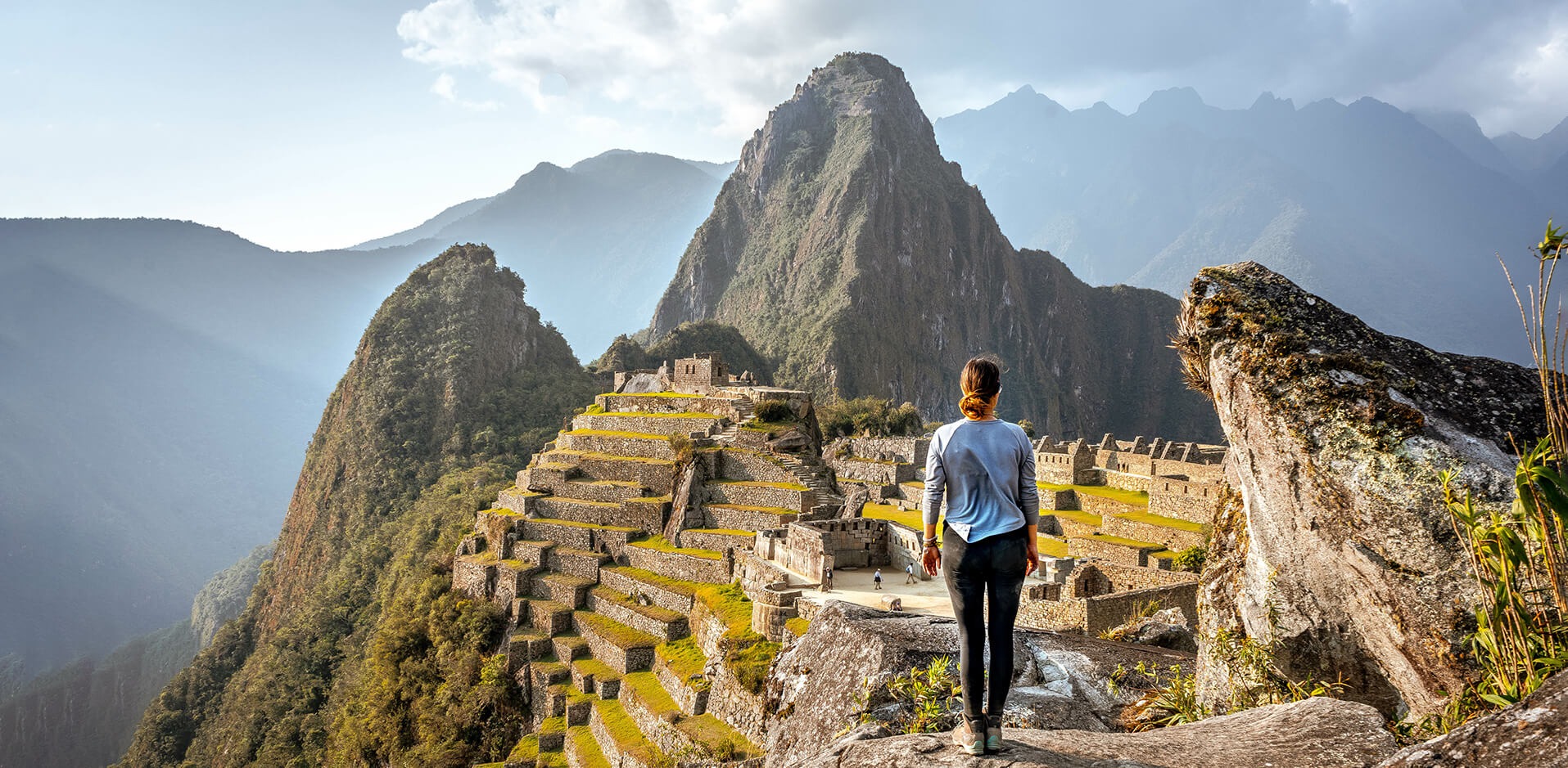 A hiker looks across Machu Picchu's stone terraces and green mountains under a bright sky in Peru.