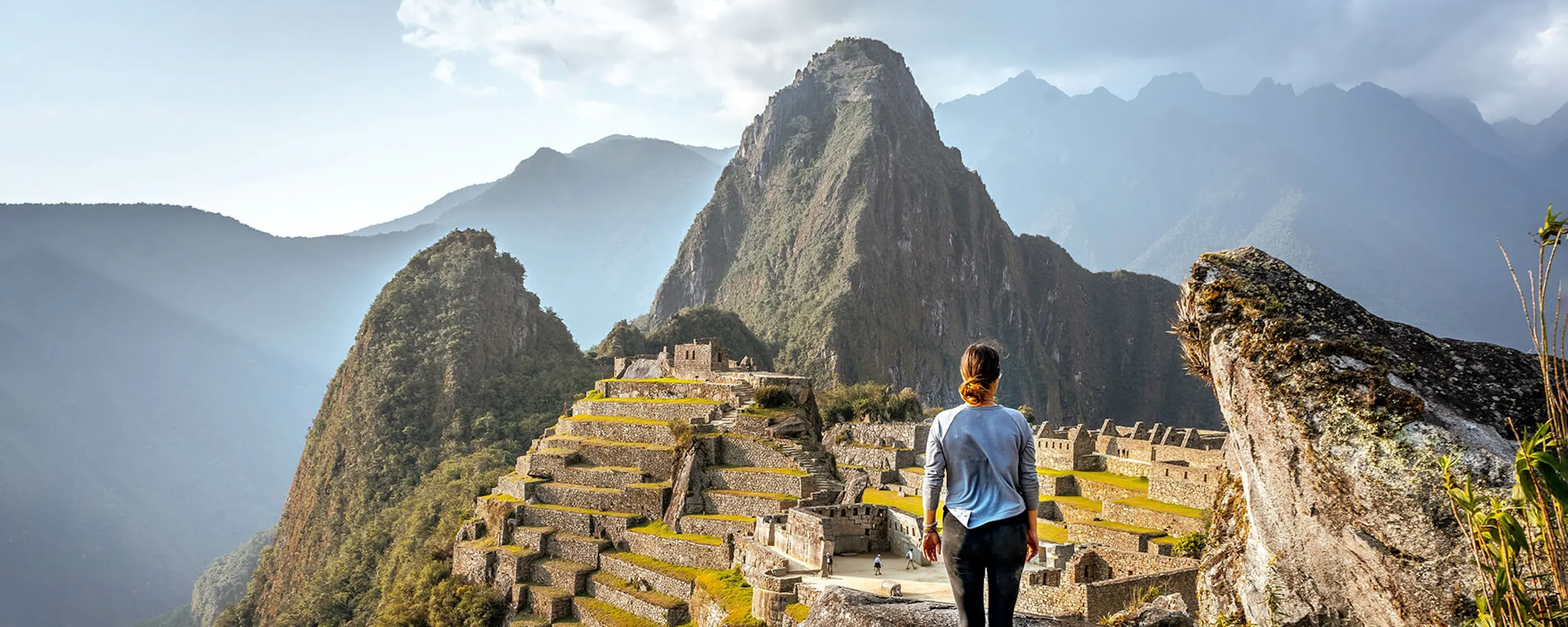 A hiker looks across Machu Picchu's stone terraces and green mountains under a bright sky in Peru.