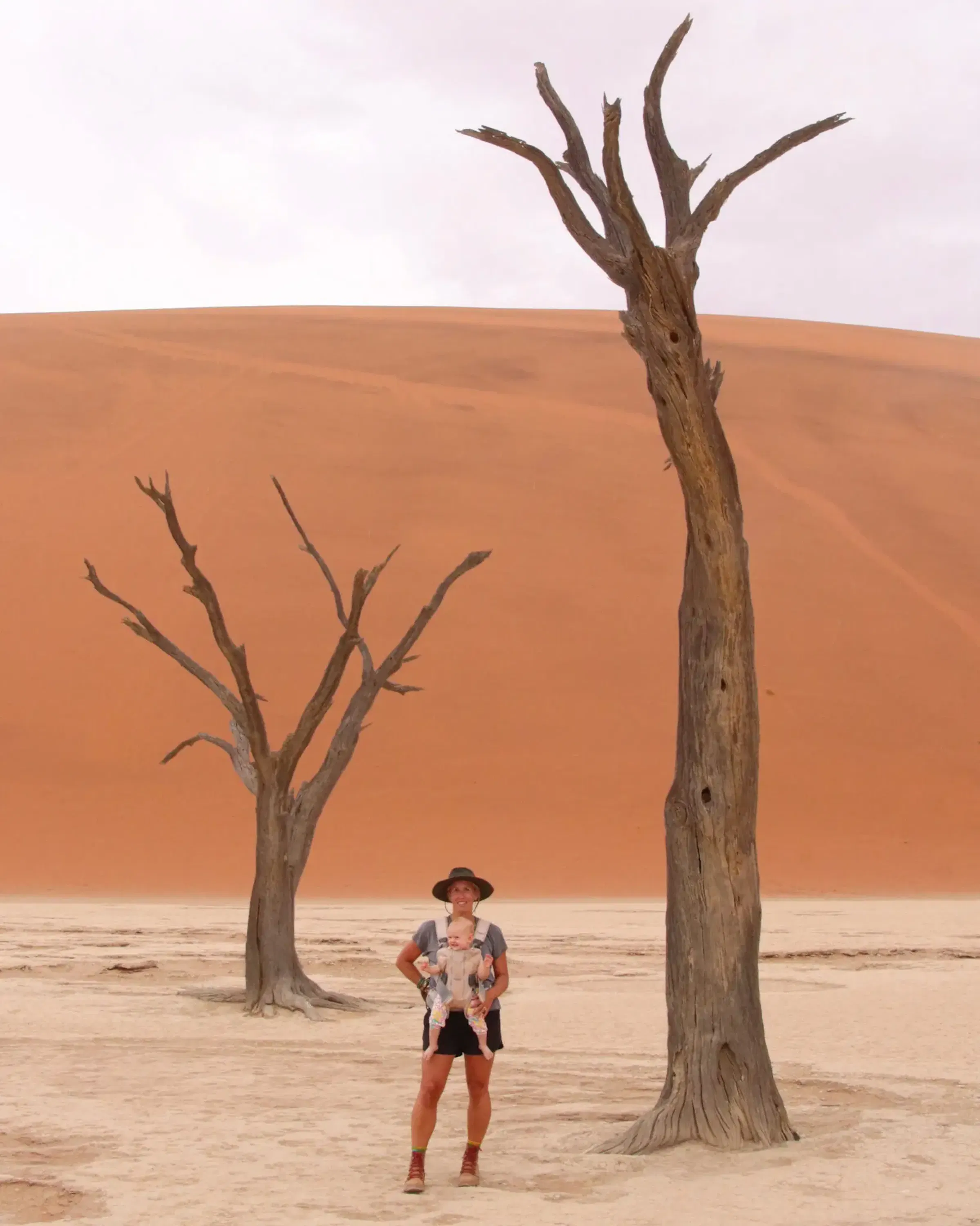 A woman with an infant strapped to her chest stands in front of a large sand dune in Namibia, with two bare trees in the background.